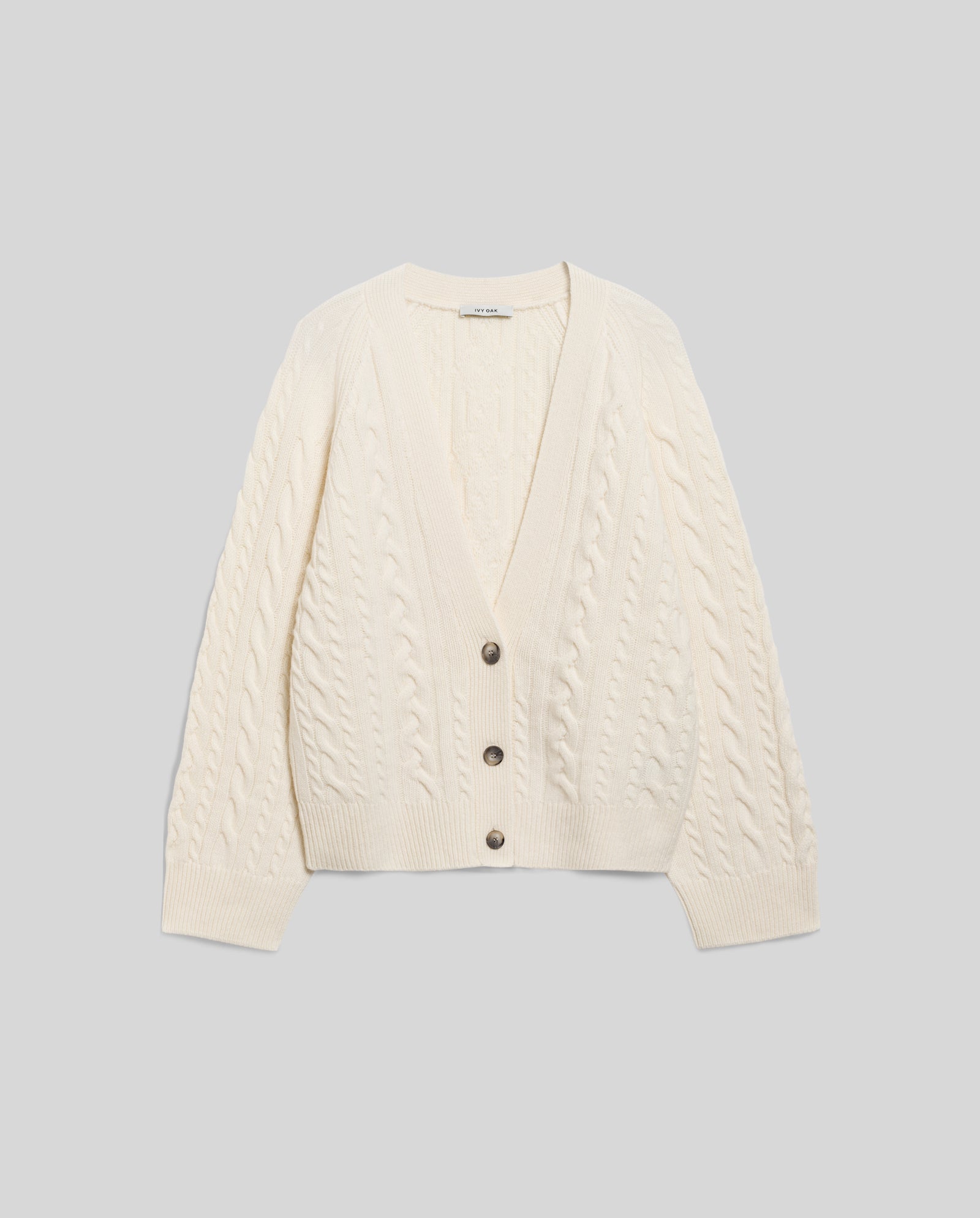 The KYMBERLY Cardigan by IVY OAK is a cream v-neck style with wide raglan sleeves, classic cable knit pattern, three dark buttons, and long sleeves, shown laid flat on a light background.