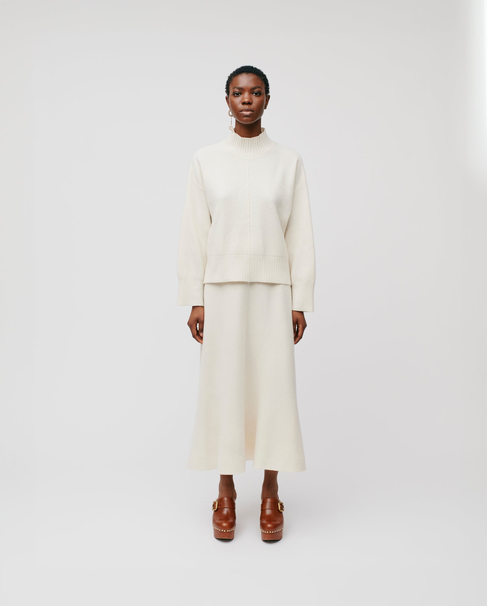 A woman wears the IVY OAK KAREEN ROSE Jumper, a cream Merino wool sweater with a stand-up collar, paired with a matching skirt and brown platform shoes against a plain white background.