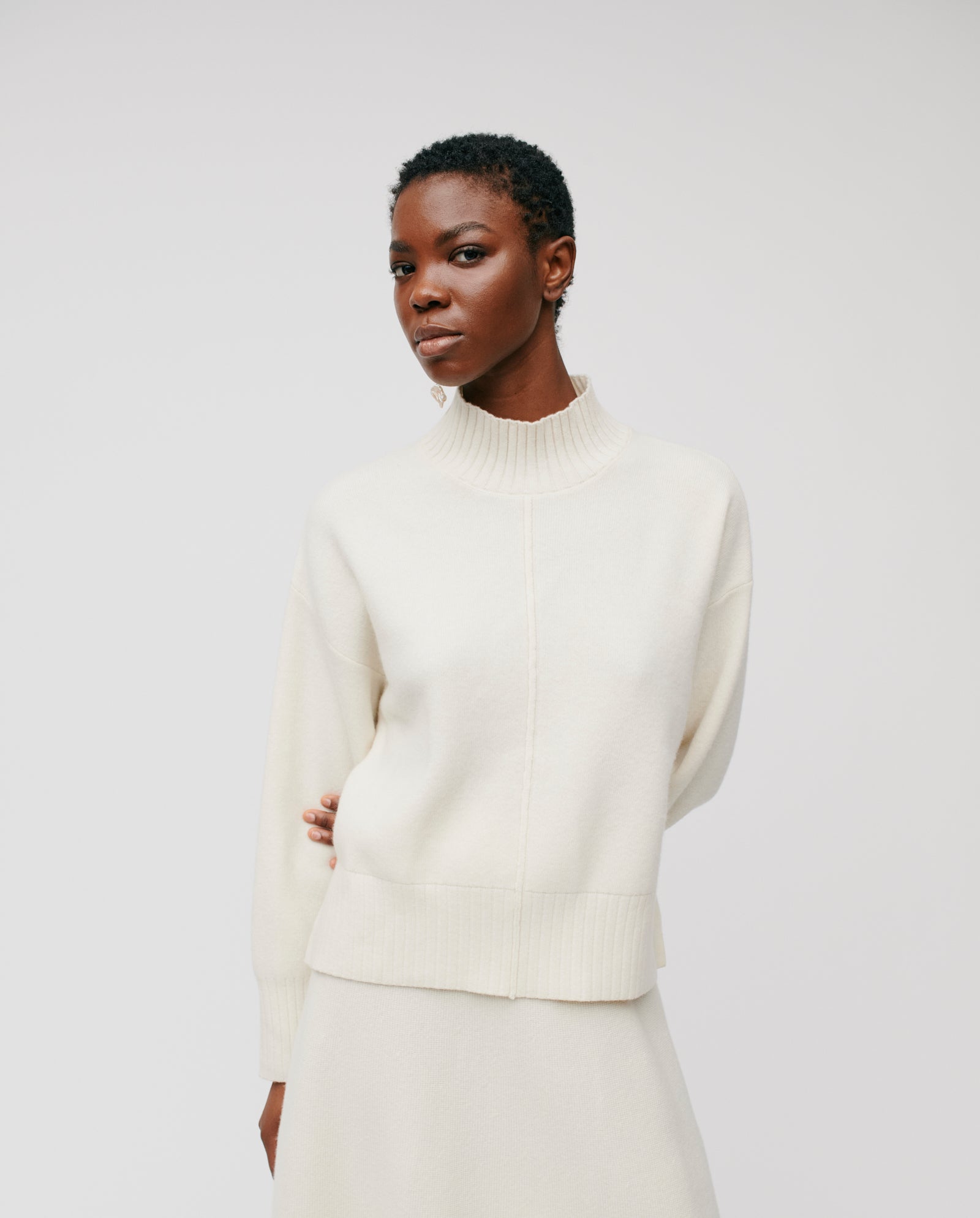 A person wears the IVY OAK KAREEN ROSE Jumper, a cream-colored Merino wool sweater with a stand-up collar, paired with a skirt against a plain white background.