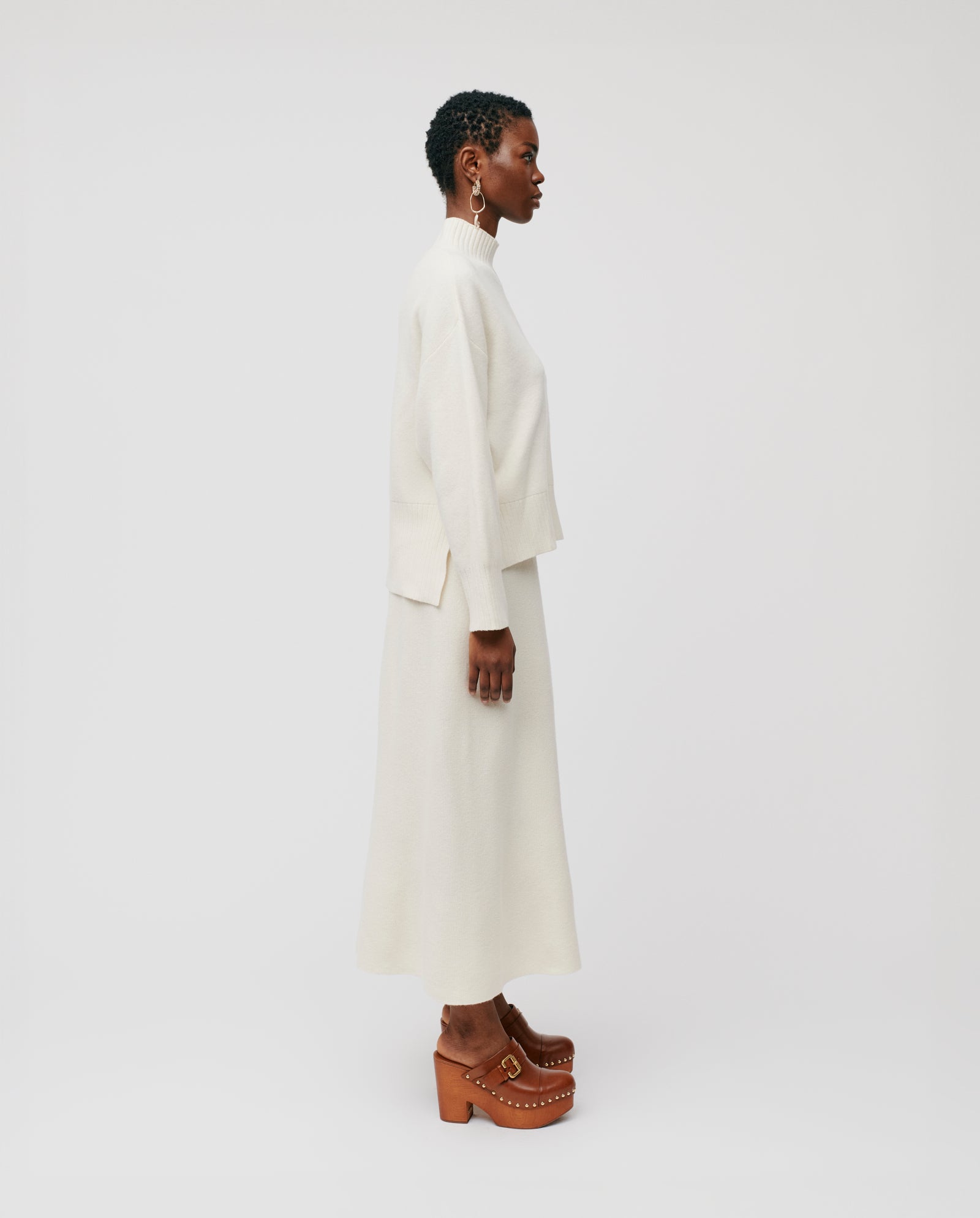 A woman wears the IVY OAK KAREEN ROSE Jumper, a cream-colored merino wool sweater, paired with a skirt and brown platform clogs, standing in profile against a white background.