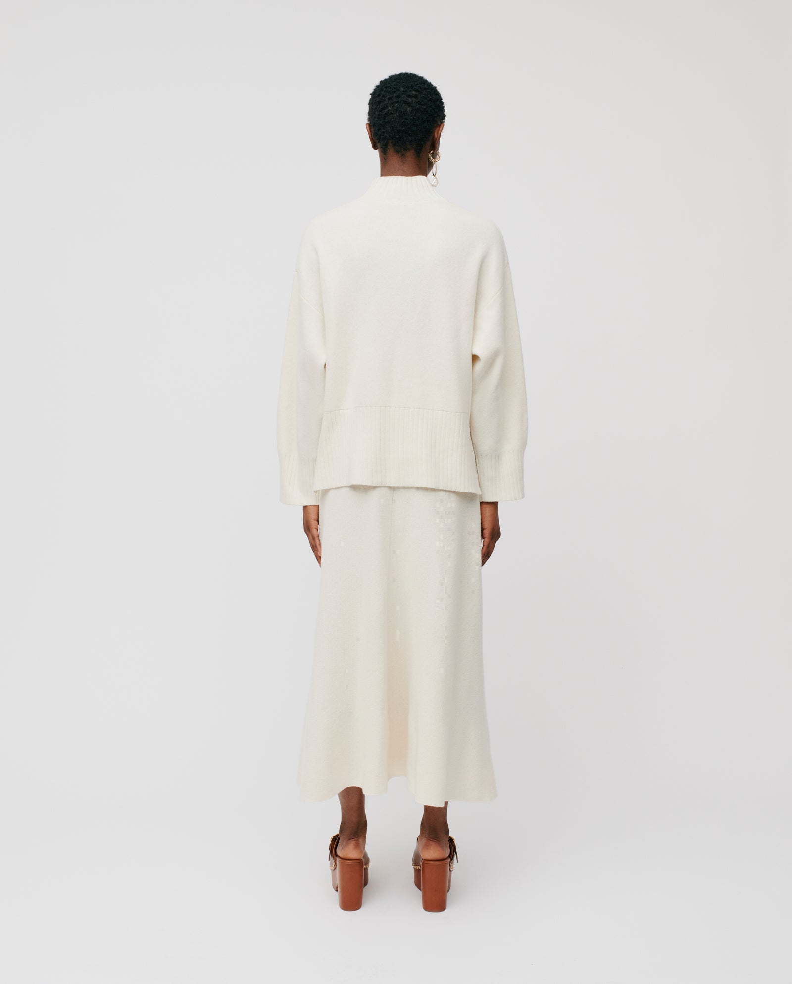 A person faces away in the IVY OAK KAREEN ROSE Jumper—a cream Merino wool sweater with a stand-up collar—styled with a skirt and brown platform heels against a plain backdrop.