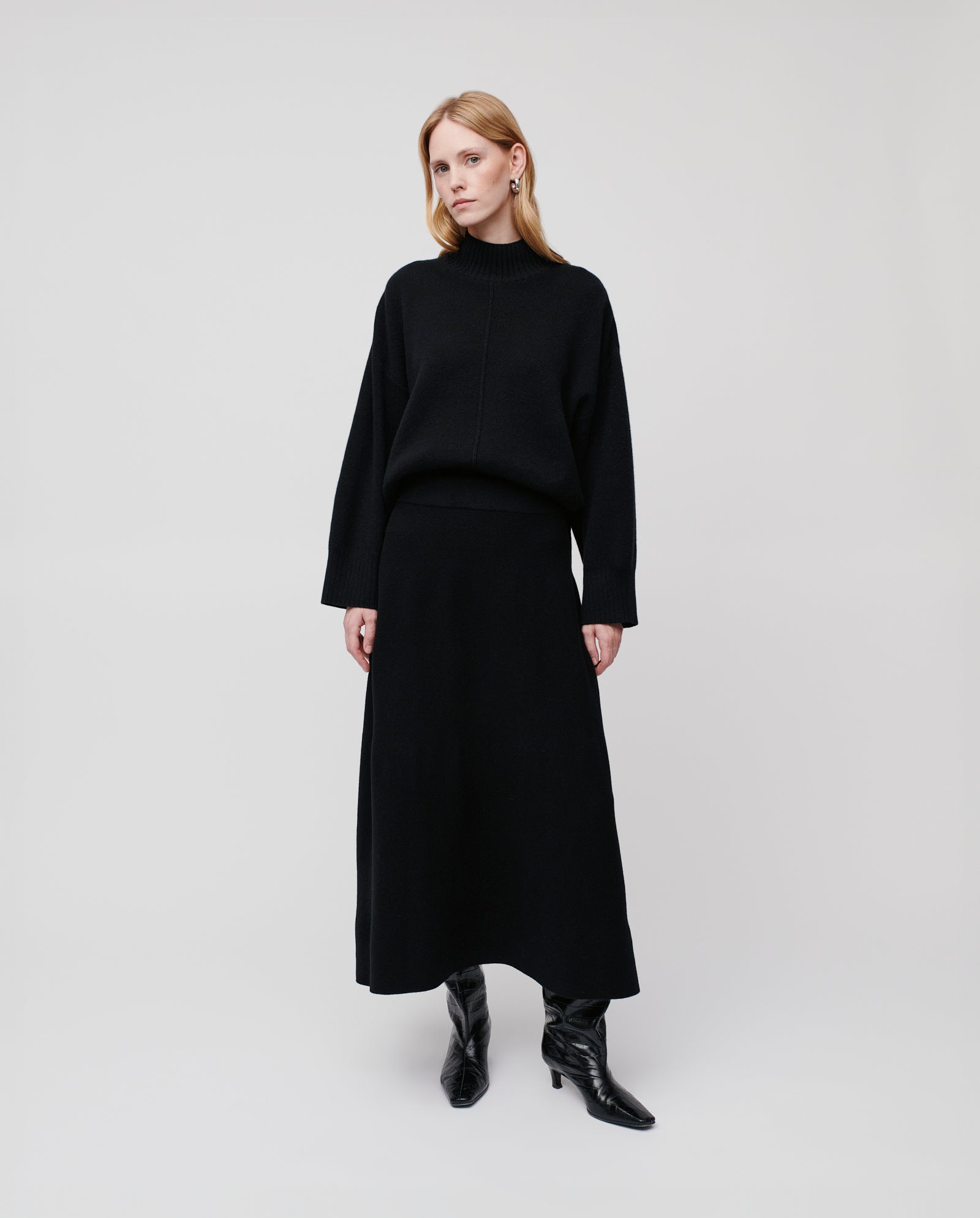 A woman wears the IVY OAK KAIA ROSE Skirt in black merino wool with an A-line silhouette, styled with a black turtleneck sweater and black boots against a white background.
