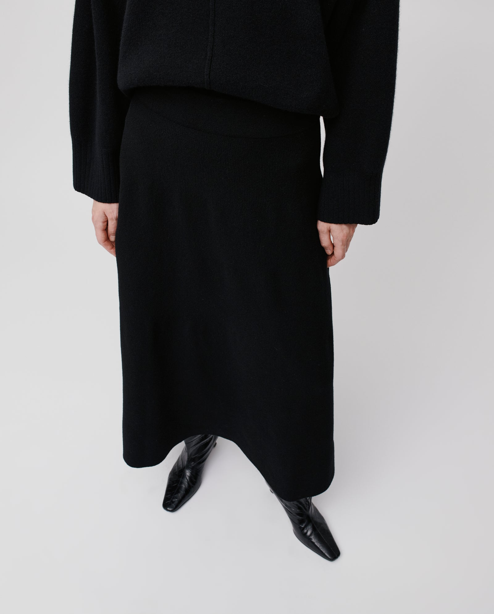 Model wears the IVY OAK KAIA ROSE Skirt—black merino wool with an A-line silhouette—paired with a black sweater and pointed boots, against a white background.