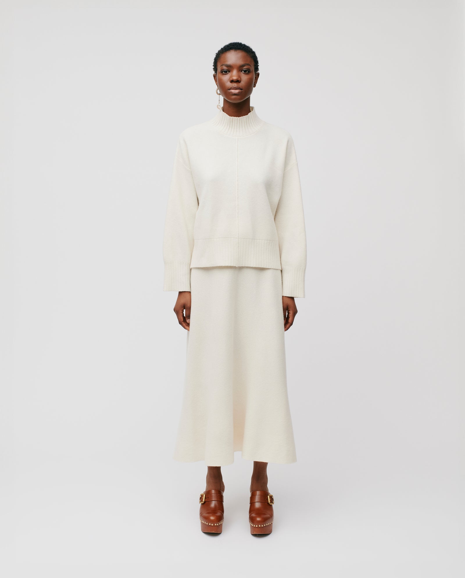 A woman in an ivory merino wool sweater and the IVY OAK KAIA ROSE Skirt with an A-line silhouette, paired with brown platform shoes, stands against a white background.
