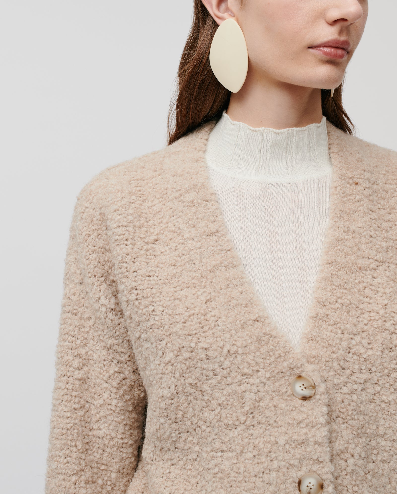 A woman models the KRISTAL Cardigan by IVY OAK, a textured beige bouclé coat, paired with a white top and large oval cream earrings against a plain background for a casual modern style.