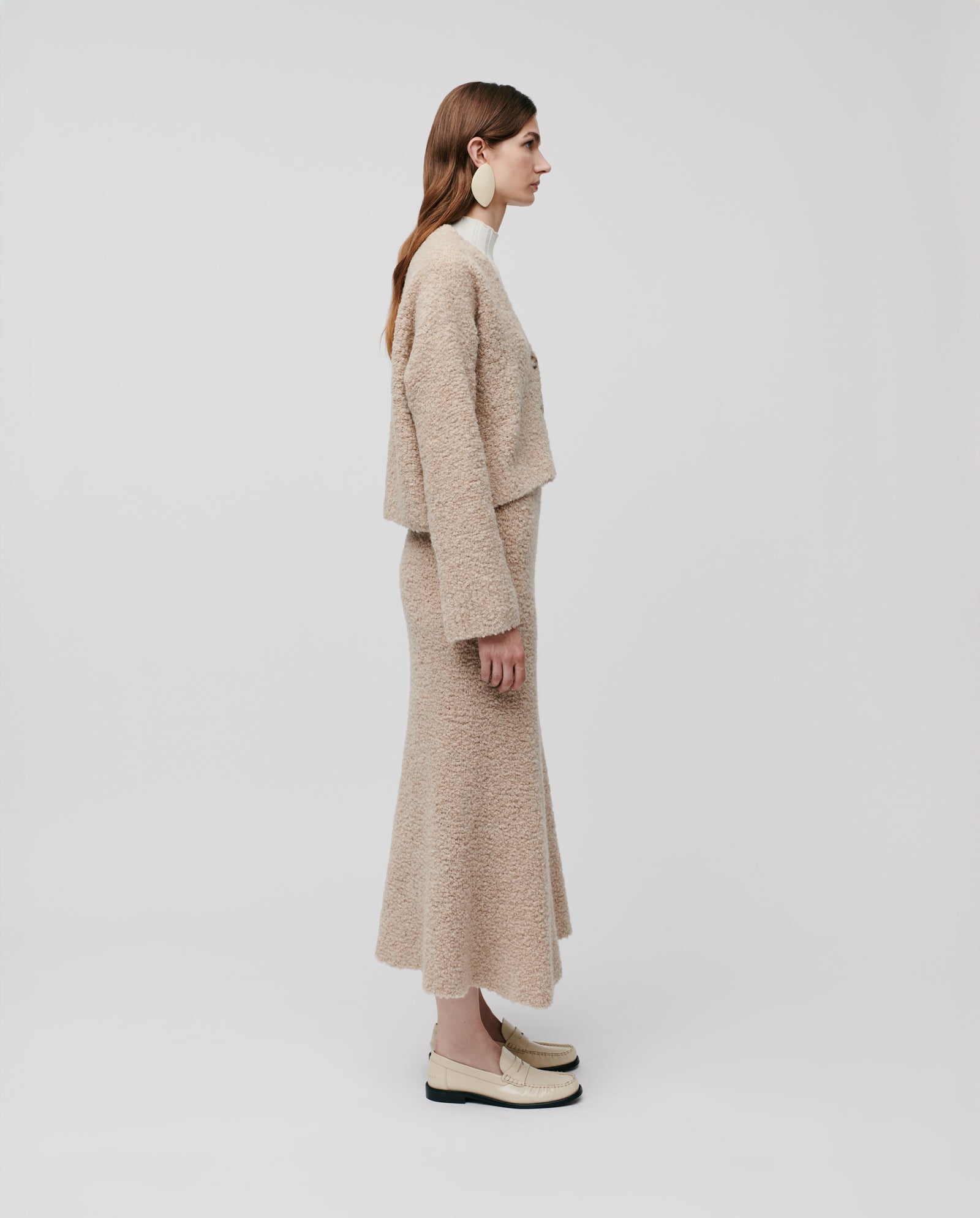 Woman in a beige bouclé yarn outfit and loafers stands in profile against a white background, highlighting the cozy KRISTAL Cardigan by IVY OAK for a casual, modern look.