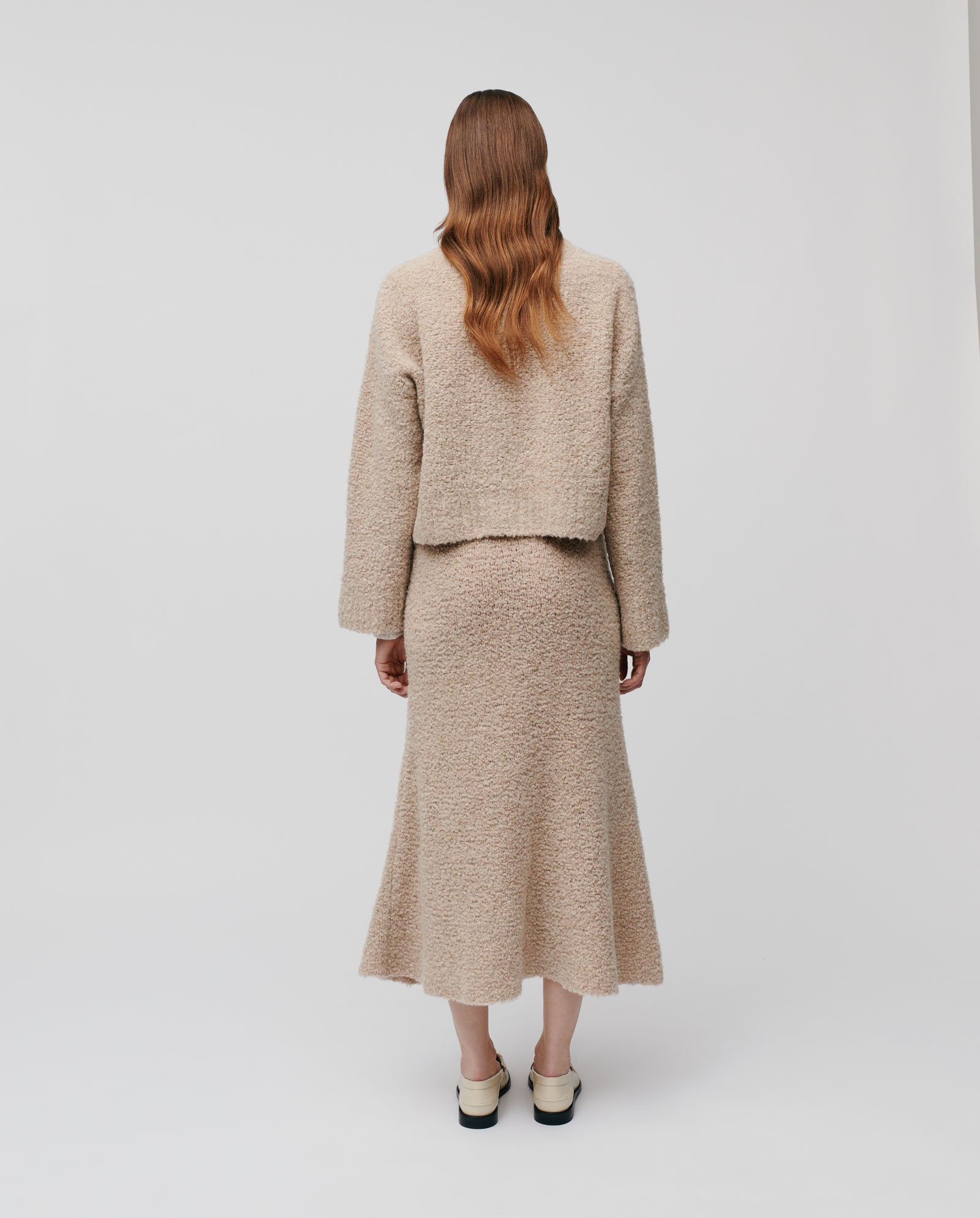 A person with long wavy hair wears the IVY OAK KRISTAL Cardigan in beige bouclé yarn, standing with their back to a plain white background for a casual, modern look.