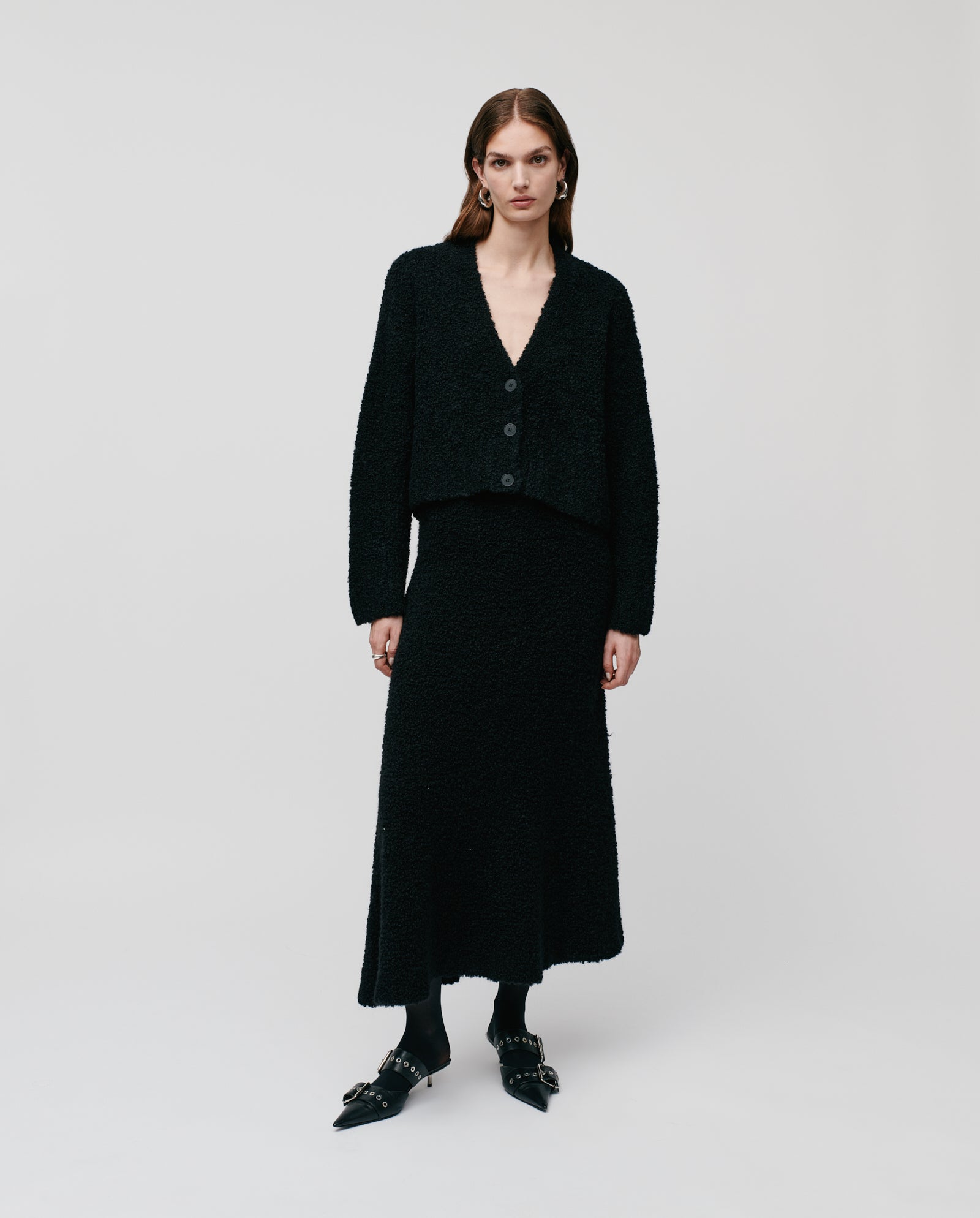 A woman wears the IVY OAK KRISTAL Cardigan, crafted from ultra soft bouclé yarn in black with a deep V-neck and wide sleeves, paired with a matching long skirt, standing against a plain white background.