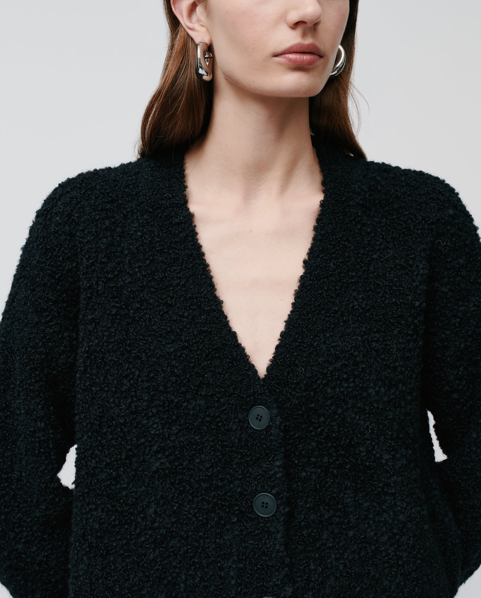 Woman wearing the KRISTAL Cardigan by IVY OAK, crafted from ultra soft bouclé yarn in black, styled with silver hoop earrings and shown from shoulders up.
