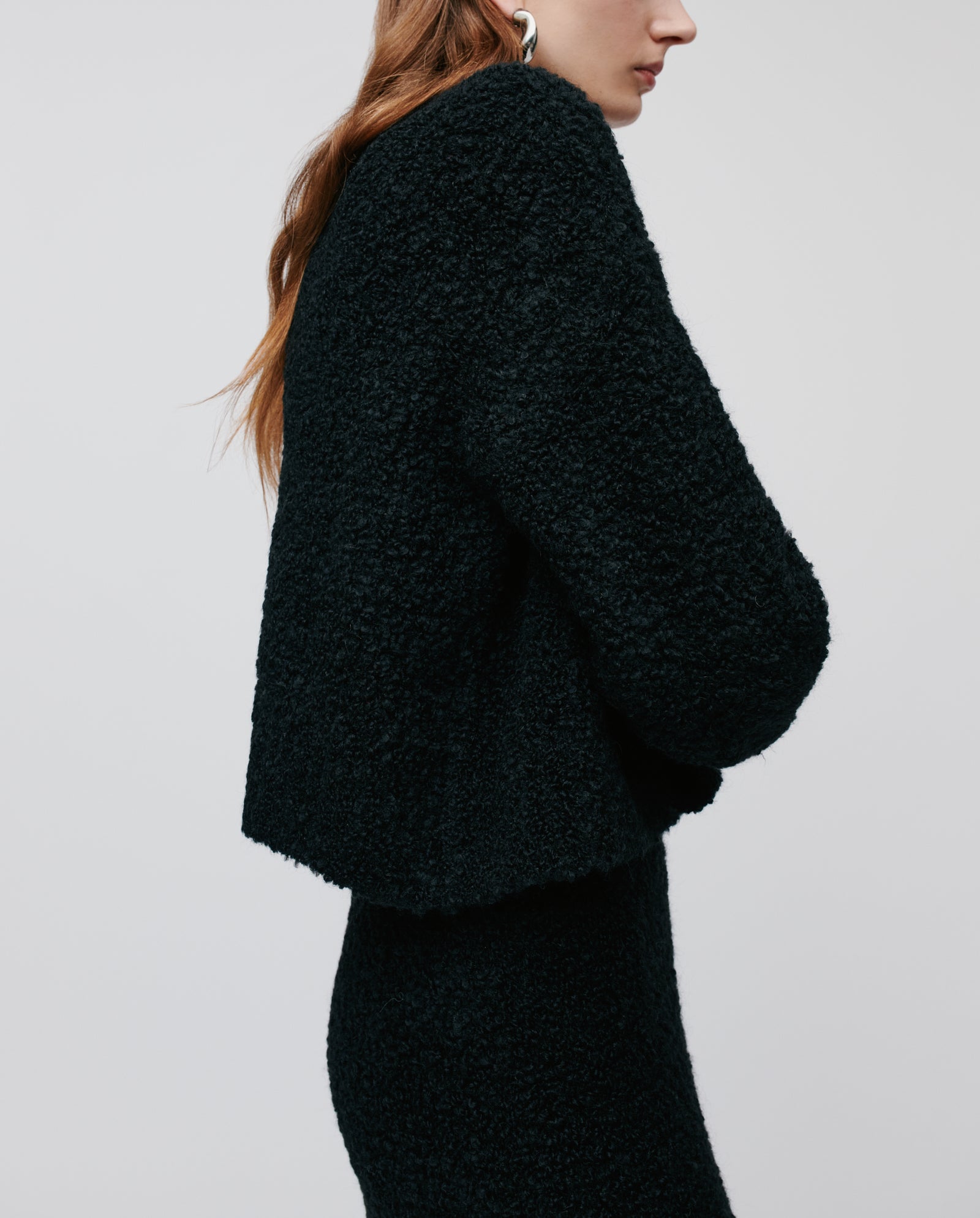 Woman in the KRISTAL Cardigan by IVY OAK—a textured black jacket with wide sleeves—paired with a skirt, shown from the side against a plain light background.