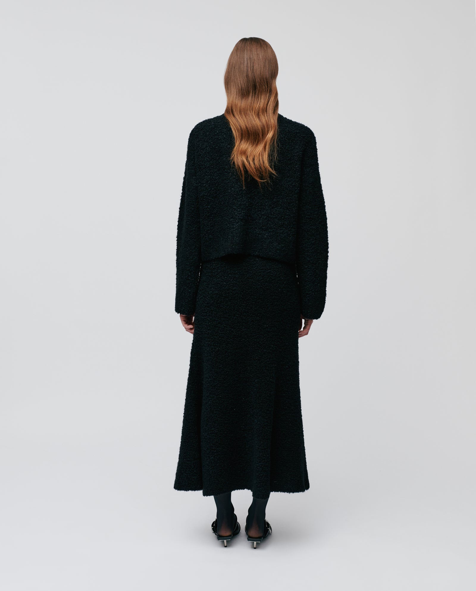 A person with long wavy hair, facing away, wears the IVY OAK KRISTAL Cardigan in black bouclé yarn with wide sleeves, paired with a textured skirt.