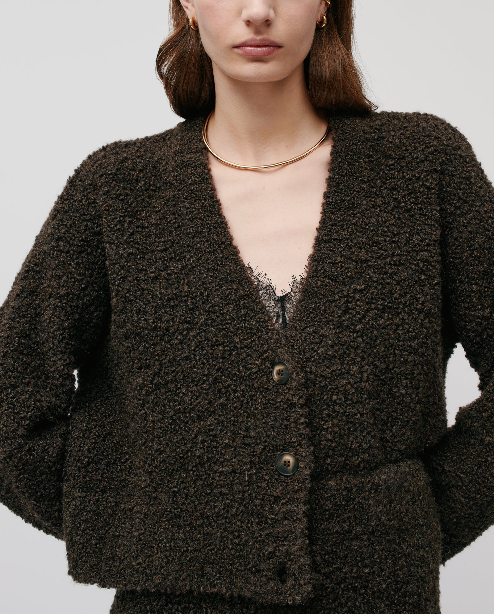A woman faces forward in a modern, casual look, wearing the IVY OAK KRISTAL Cardigan—an ultra soft bouclé yarn V-neck with large buttons in textured dark brown—paired with a gold necklace.