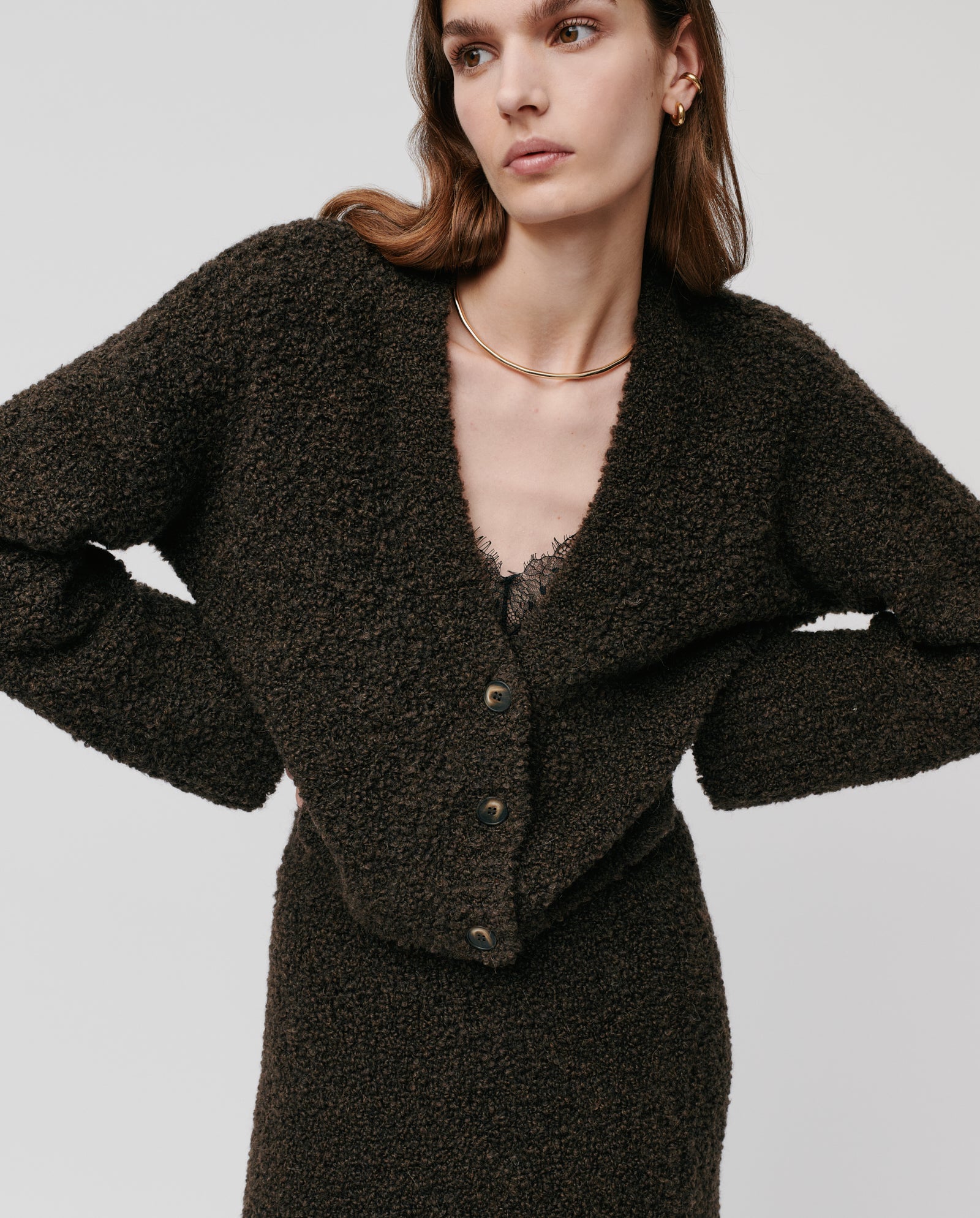 A woman models the IVY OAK KRISTAL Cardigan, made from ultra-soft bouclé yarn, paired with a matching skirt. She stands with hands on hips against a plain background, showcasing a chic and modern look.