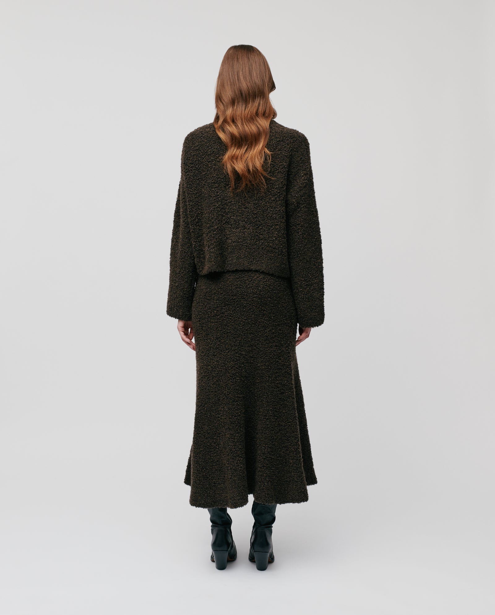 A woman with wavy brown hair models the IVY OAK KRISTAL Cardigan, a modern, ultra-soft bouclé yarn piece in a dark hue, standing with her back to the camera.