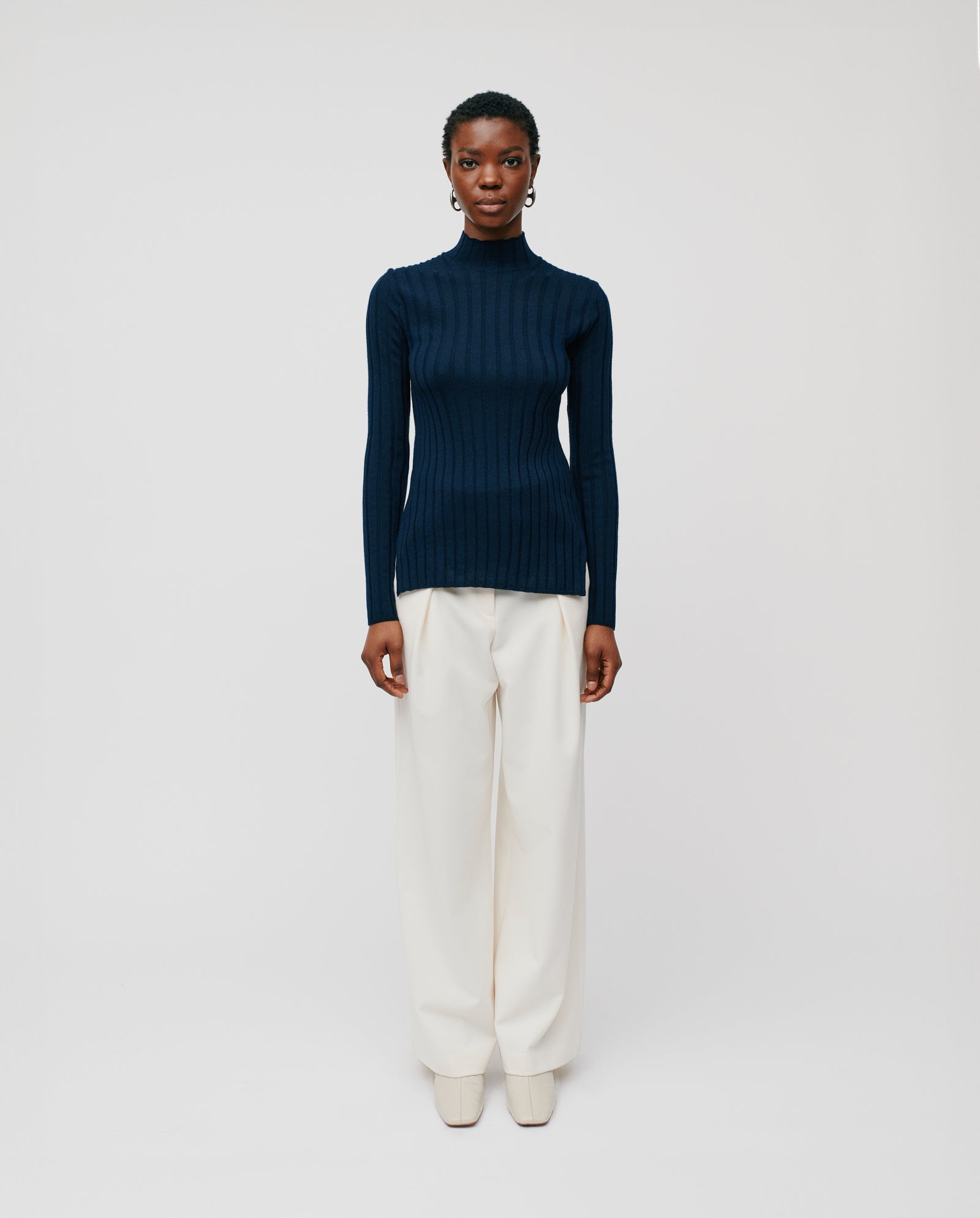 A woman in the IVY OAK KACY ROSE Jumper—a navy ribbed merino wool sweater—and wide white trousers stands against a plain light background.