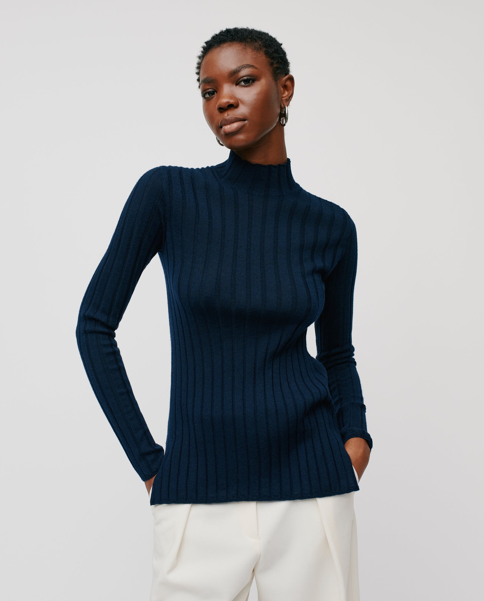 Model wears the IVY OAK KACY ROSE Jumper, a fitted ribbed navy turtleneck, paired with white pants and posing against a light background.