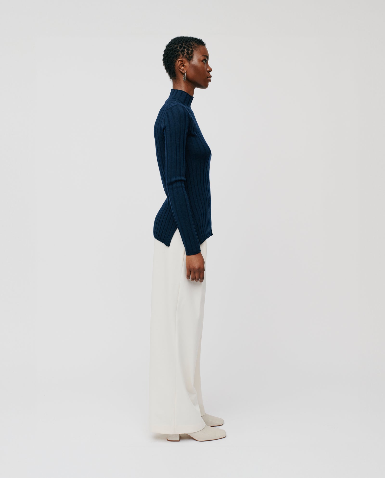 A woman stands in profile against a plain background, wearing the IVY OAK KACY ROSE Jumper in navy merino wool paired with white wide-leg pants.