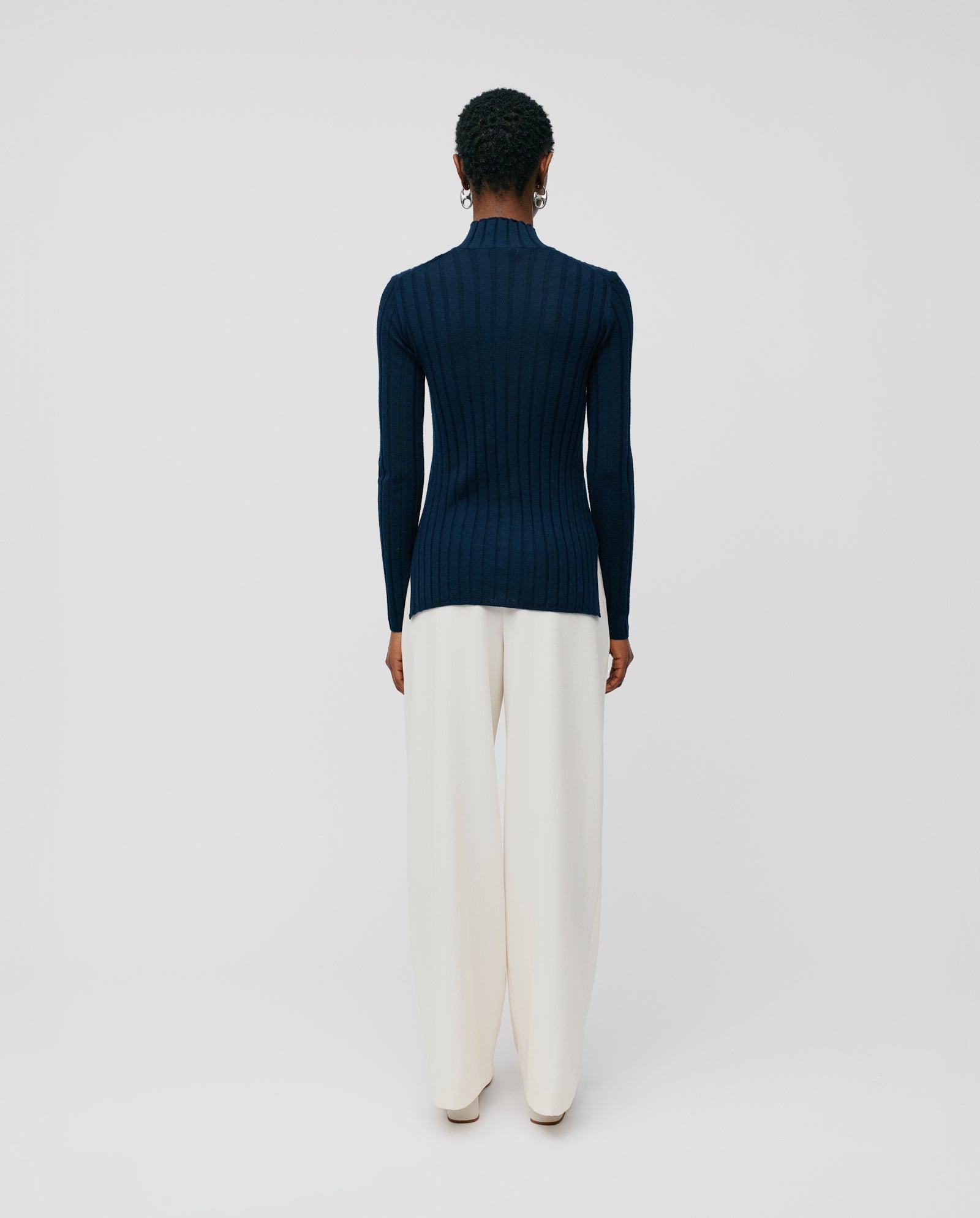A person stands facing away in the IVY OAK KACY ROSE Jumper, a navy knitted sweater with a ribbed structure, paired with wide-leg white pants against a plain background.