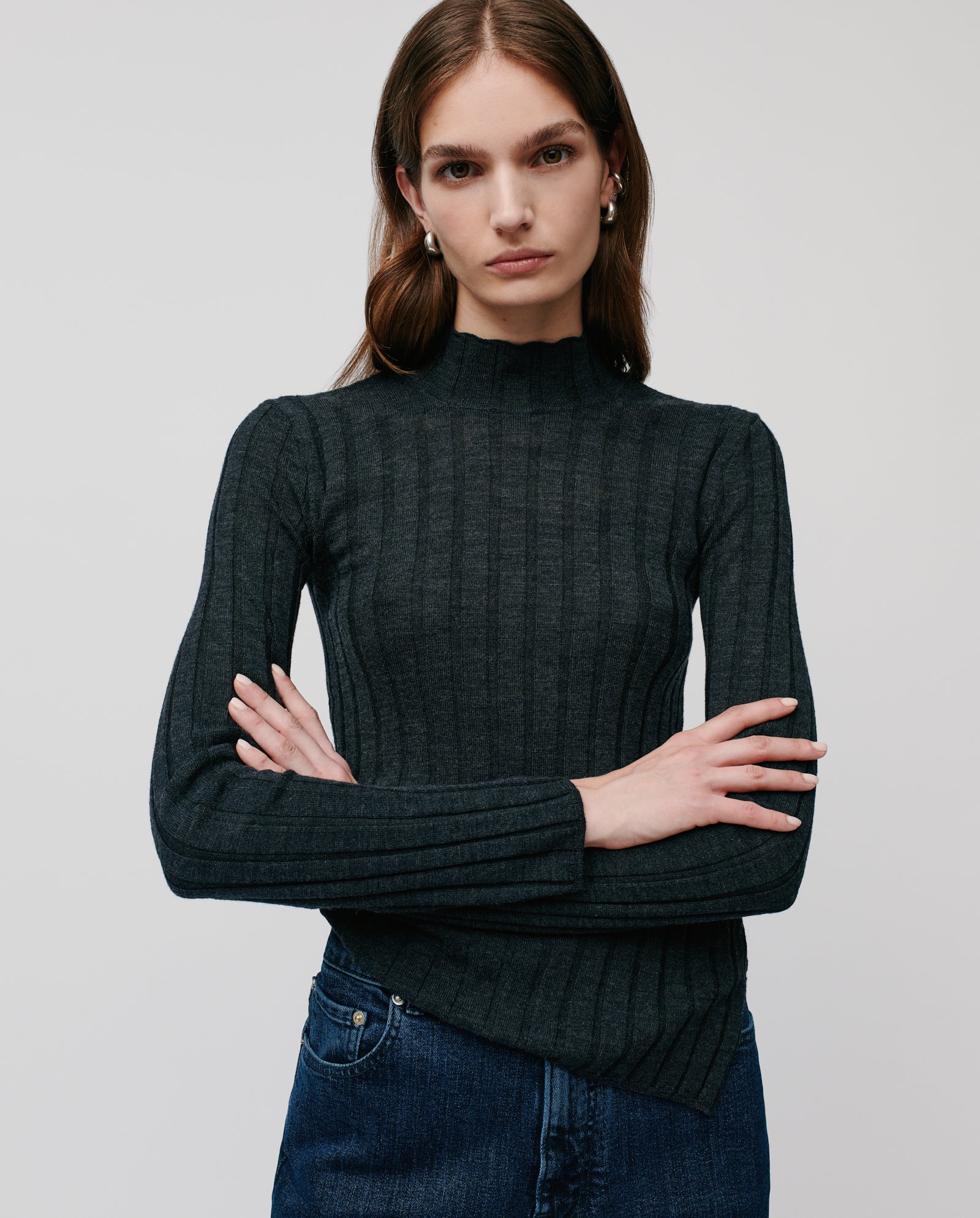 Woman with long brown hair wears the IVY OAK KACY ROSE Jumper, a dark ribbed sweater, paired with jeans, standing with arms crossed.