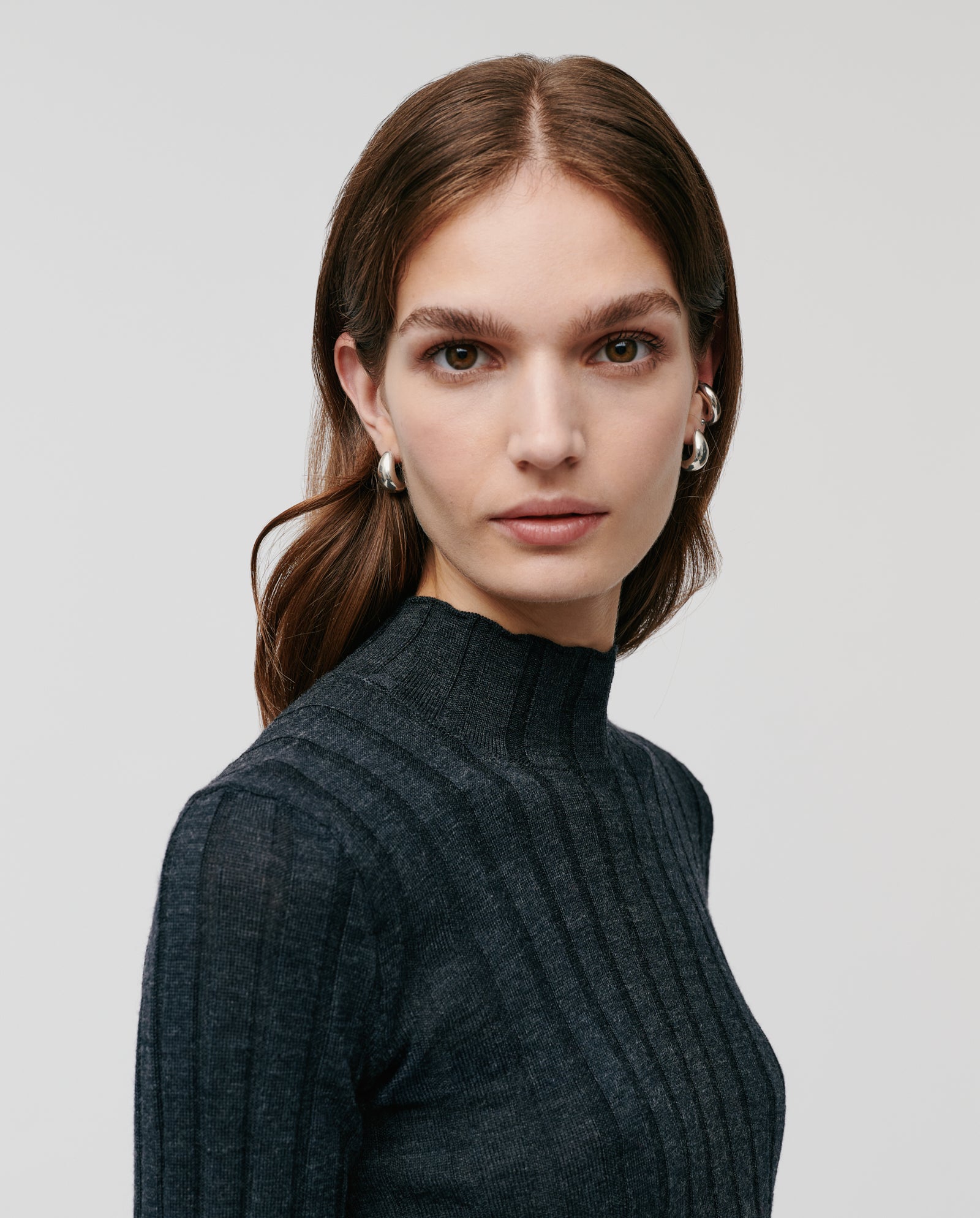 Woman with long brown hair wears the IVY OAK KACY ROSE Jumper, a dark ribbed merino wool sweater, and silver hoop earrings, looking at the camera.