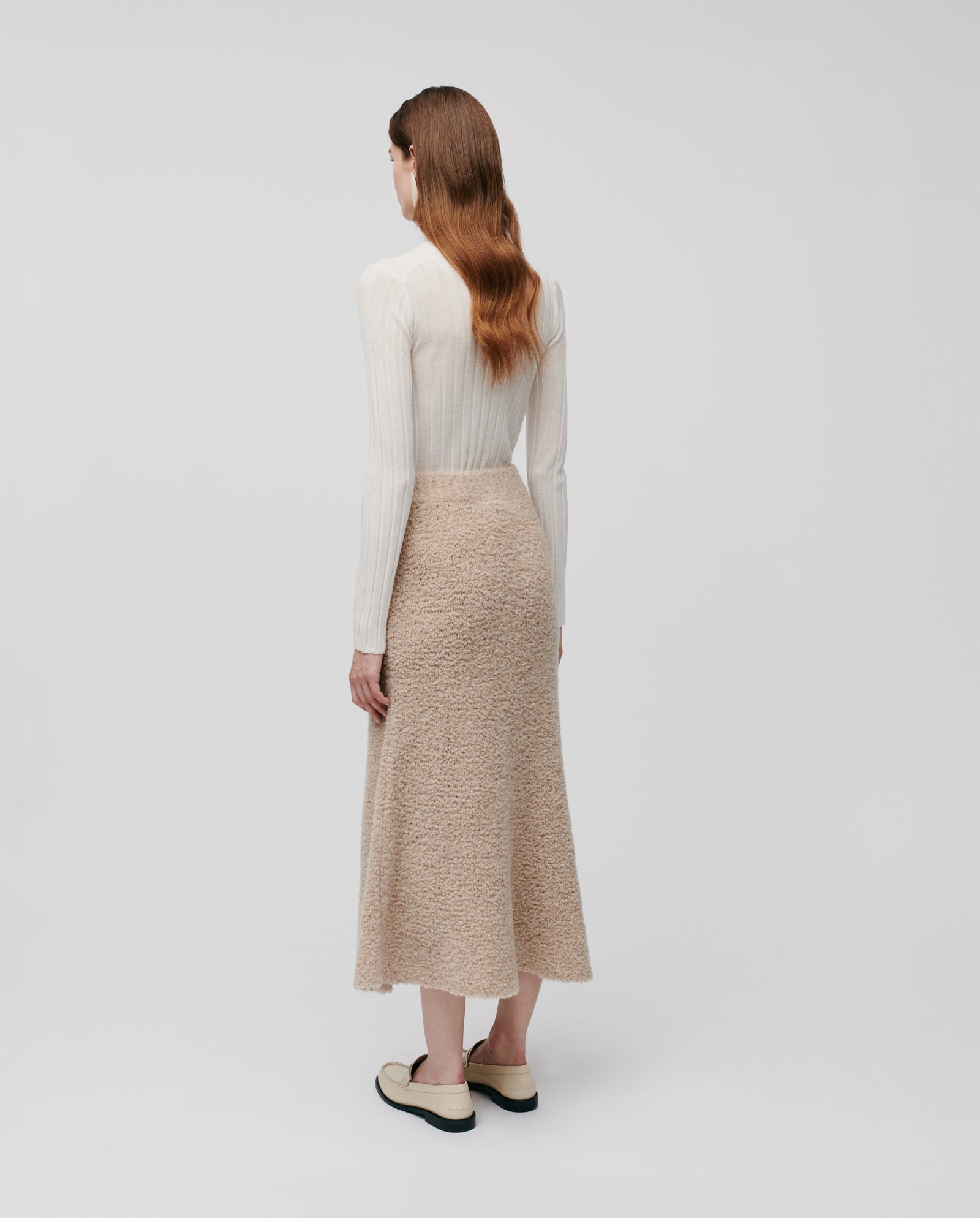 Woman with long wavy hair in a cream sweater, wearing the IVY OAK KRISTYN Skirt—an A-line beige skirt with elastic waistband—and loafers, seen from behind.