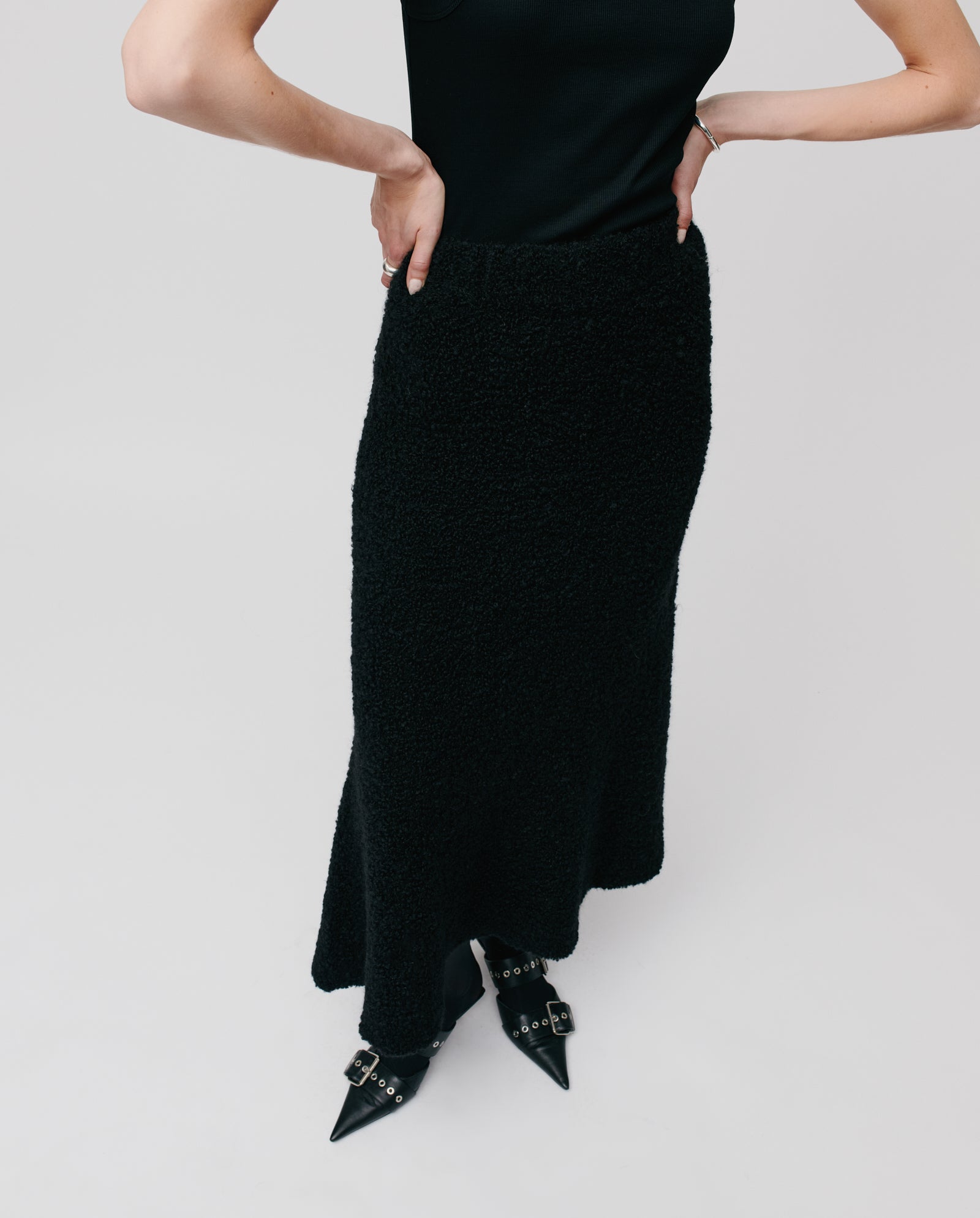 A person wearing a black sleeveless top, the IVY OAK KRISTYN Skirt in an A-line silhouette crafted from bouclé yarn, and pointed black shoes with buckles.