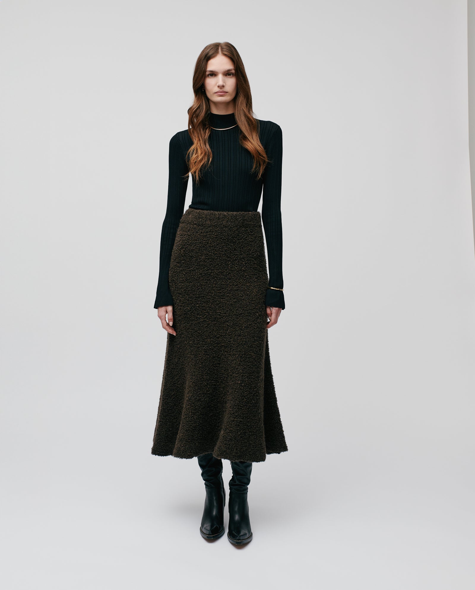 A woman stands against a plain light background wearing IVY OAK’s KRISTYN Skirt, a textured brown bouclé yarn A-line design with an elastic waistband, paired with a black long-sleeve top.