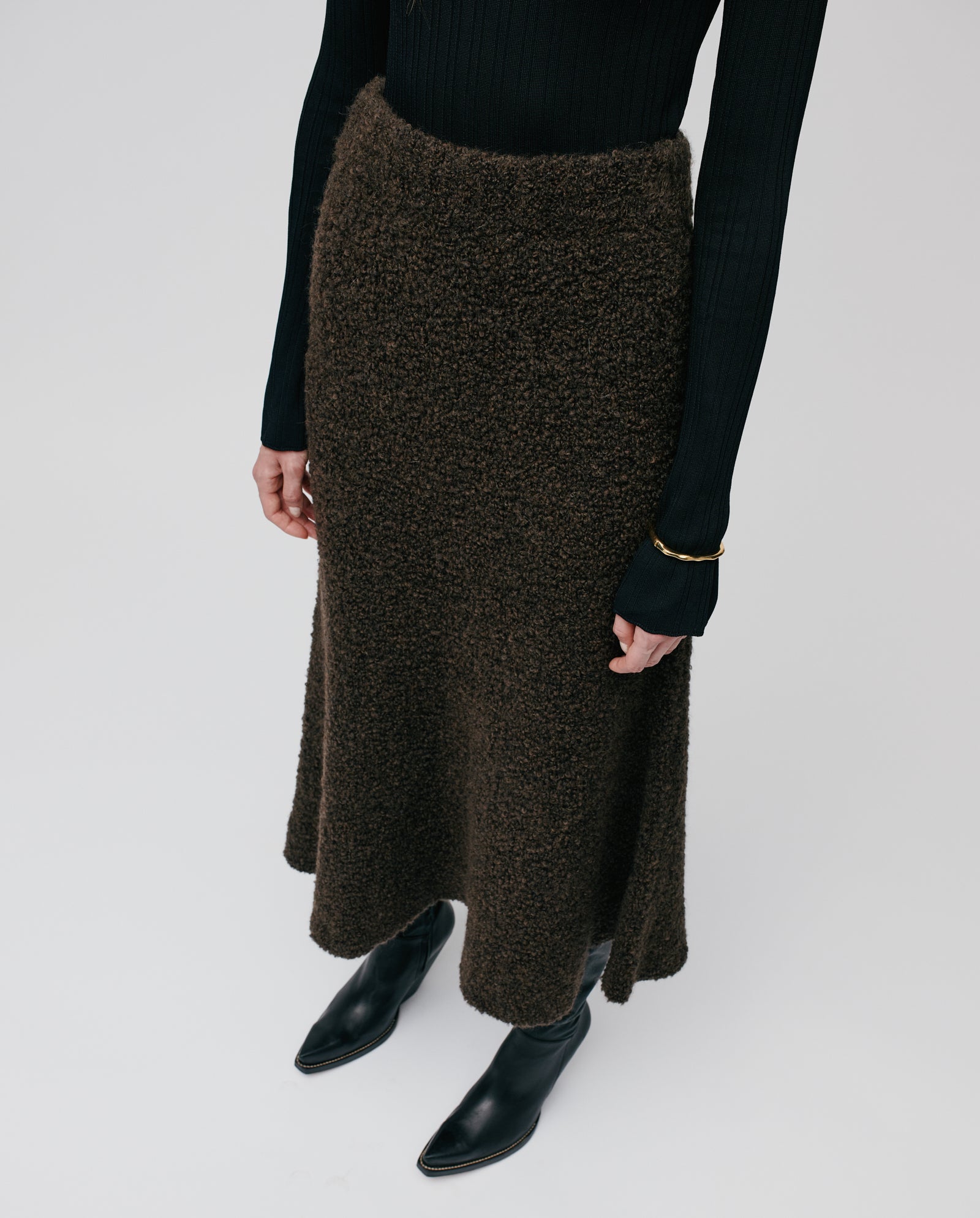 A woman wears the IVY OAK KRISTYN Skirt—a textured brown A-line skirt with an elastic waistband—paired with a black ribbed top, black boots, and a gold bracelet.