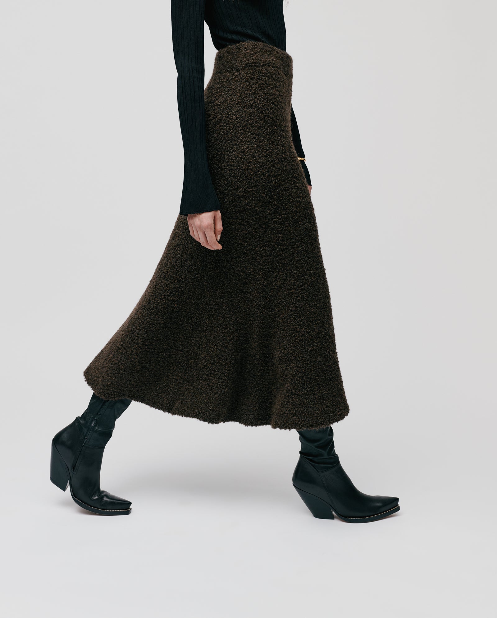 A person wearing the IVY OAK KRISTYN textured brown A-line skirt with an elastic waistband, paired with a black top and black heeled boots, walking sideways.