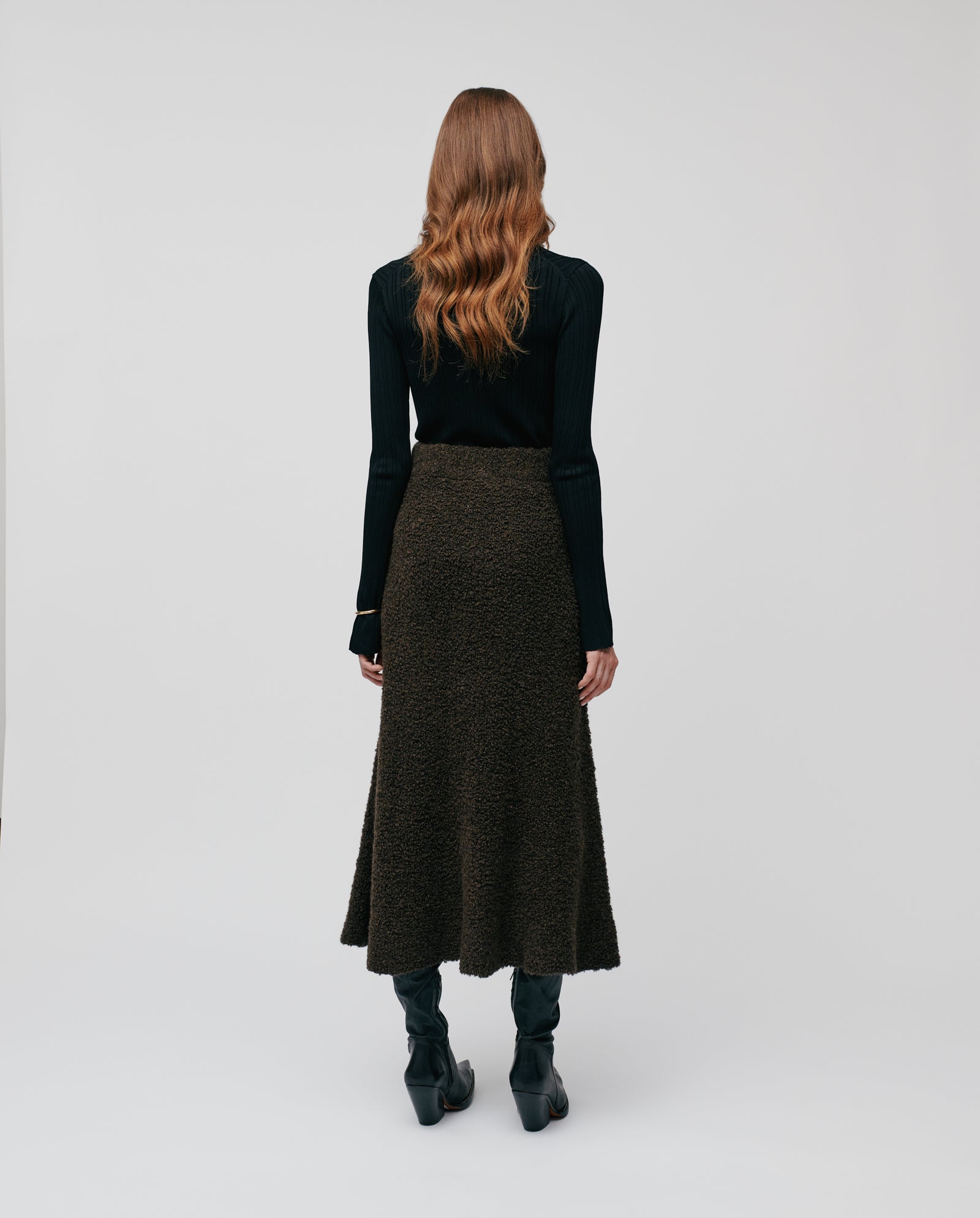 A woman with wavy brown hair, dressed in a black top and the IVY OAK KRISTYN Skirt—a long A-line skirt with an elastic waistband—stands facing away.