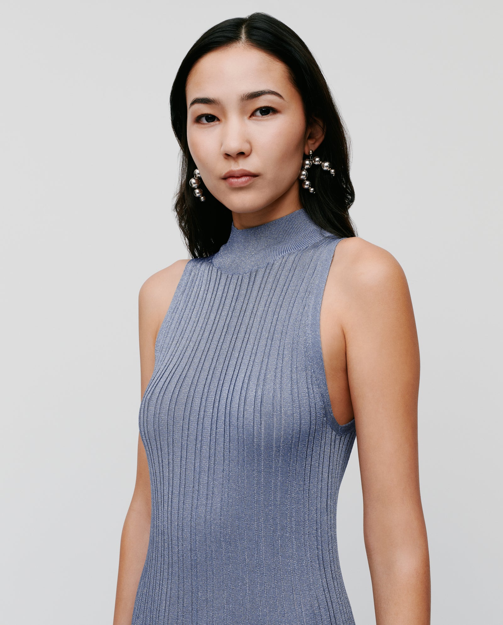 A woman in a sleeveless, fitted IVY OAK FLORA dress made of ribbed, metallic blue glitter knit, with long dark hair and statement earrings.