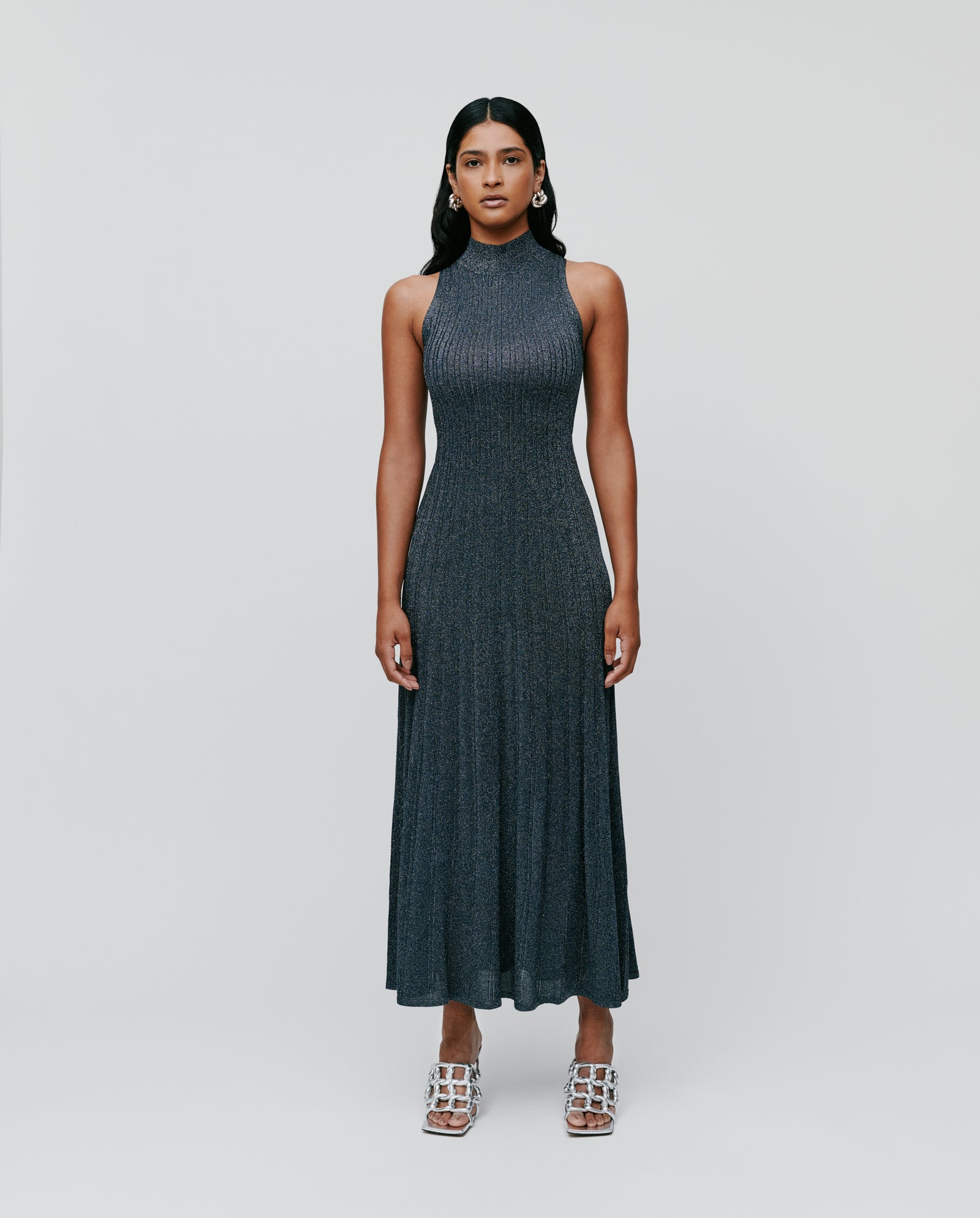 A woman in a sleeveless, high-neck, dark gray IVY OAK FLORA Dress with a glitter yarn design and silver heels stands against a plain light background.