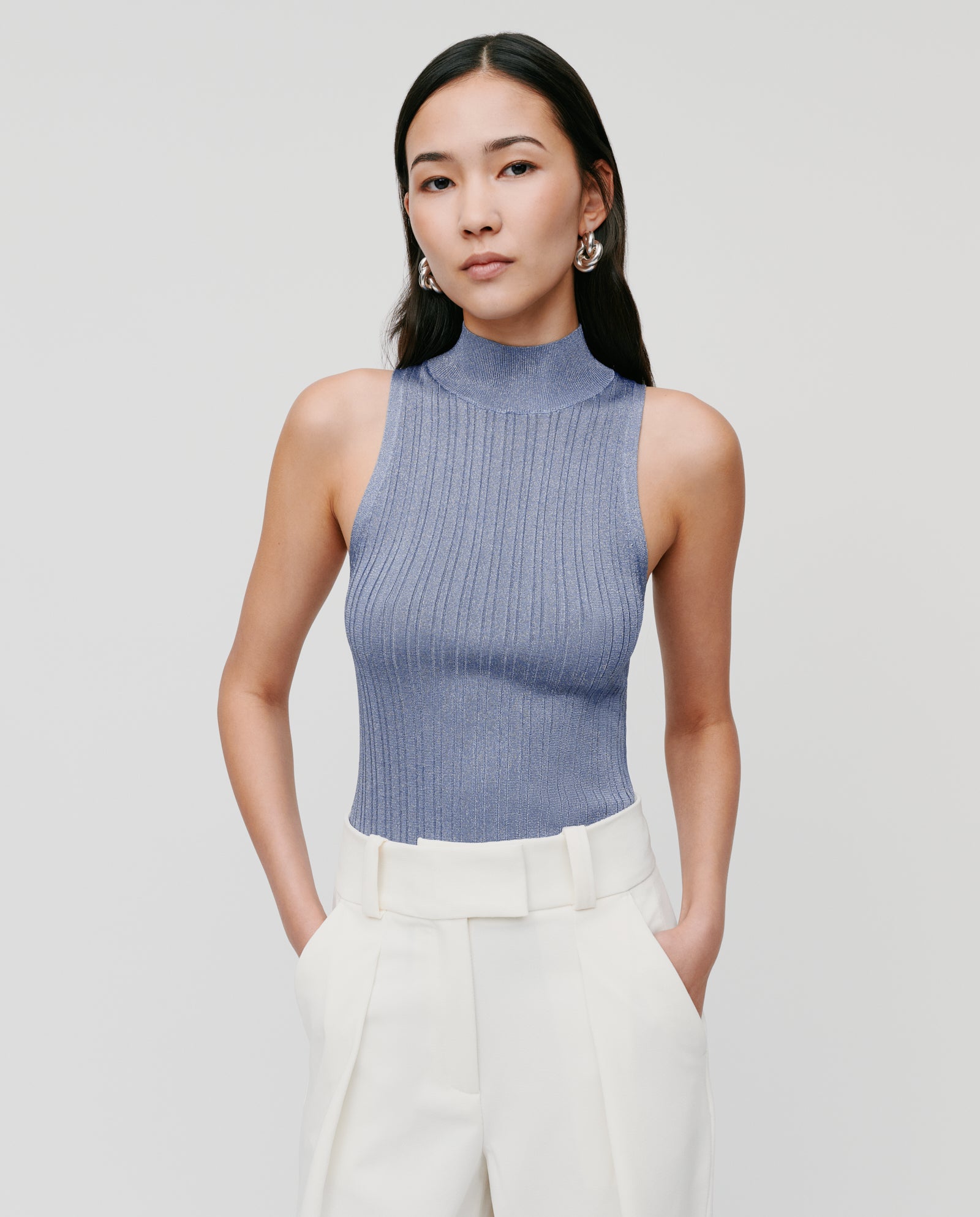 A woman wears the FINJA Top by IVY OAK, an elegant high-collar fitted gray knit, paired with high-waisted white pants, standing against a plain background.