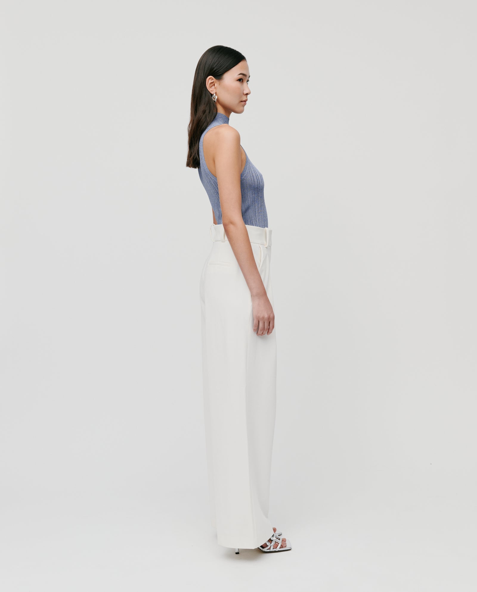 A woman in the IVY OAK FINJA Top, a blue fitted knit, and white wide-leg pants stands in profile against a white background.