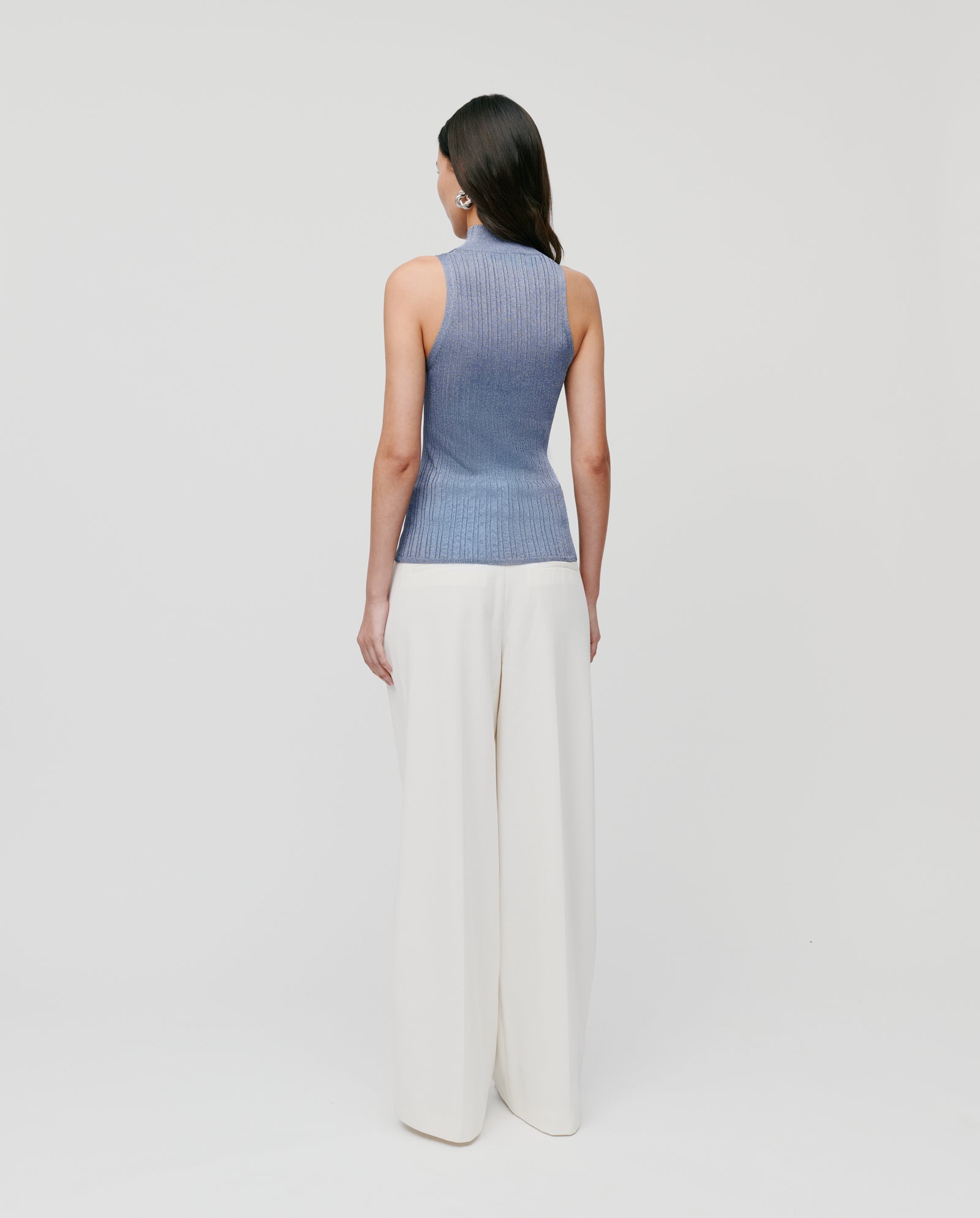 A woman stands facing away in the IVY OAK FINJA Top, a fitted knit with an elegant high collar, paired with wide-leg white pants against a plain background.