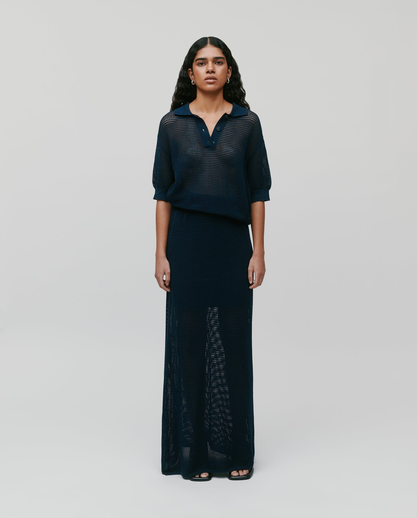 A woman stands against a plain background, wearing the IVY OAK FLO Top— a navy summer open-knit polo shirt with three buttons—paired with a matching long skirt.