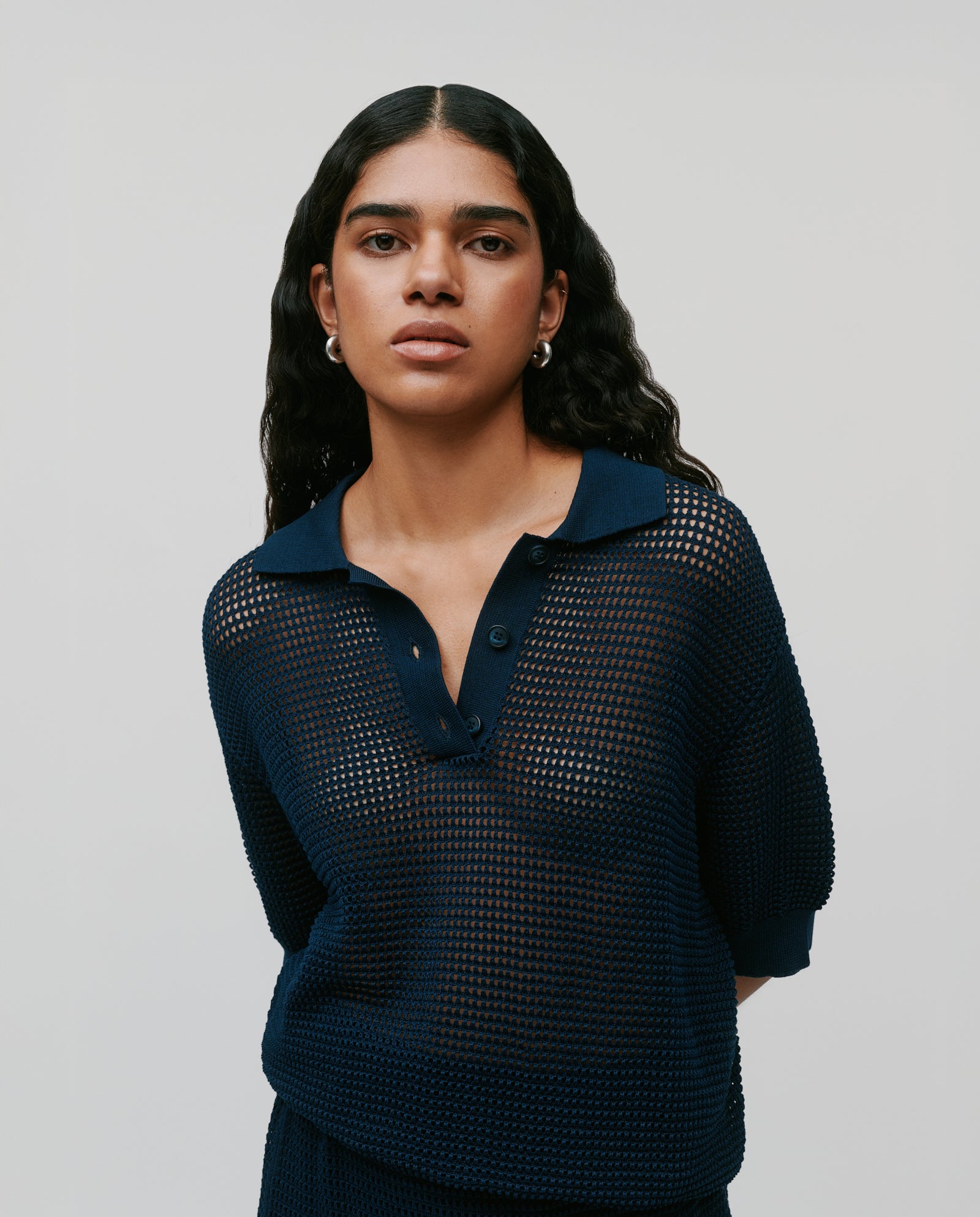 Woman with long wavy hair wears the IVY OAK FLO Top, a navy blue summer open knit sweater with three buttons, standing against a plain light background.