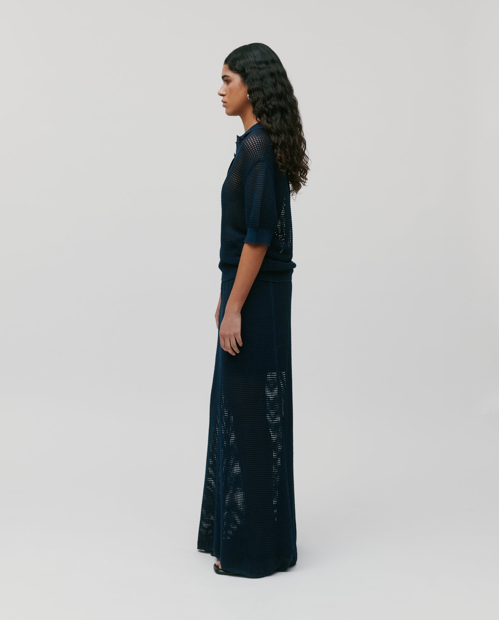 A woman in profile stands against a light background wearing the navy blue FLO Top by IVY OAK, featuring an open knit mesh, three buttons, and styled with a long skirt.