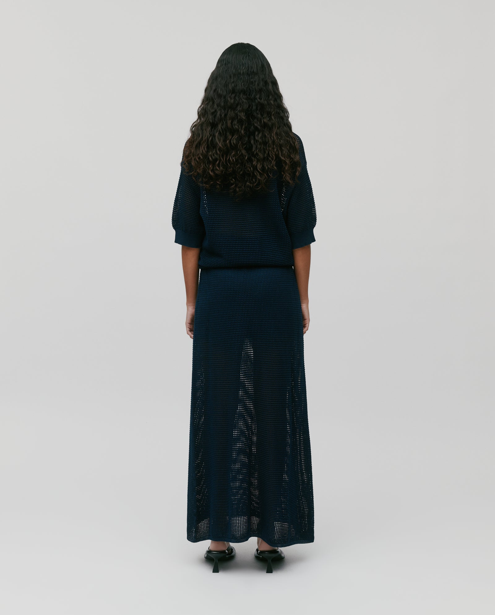 A woman with long curly hair, standing with her back to the camera, wears the FLO Top by IVY OAK—a navy summer open knit top featuring three buttons.