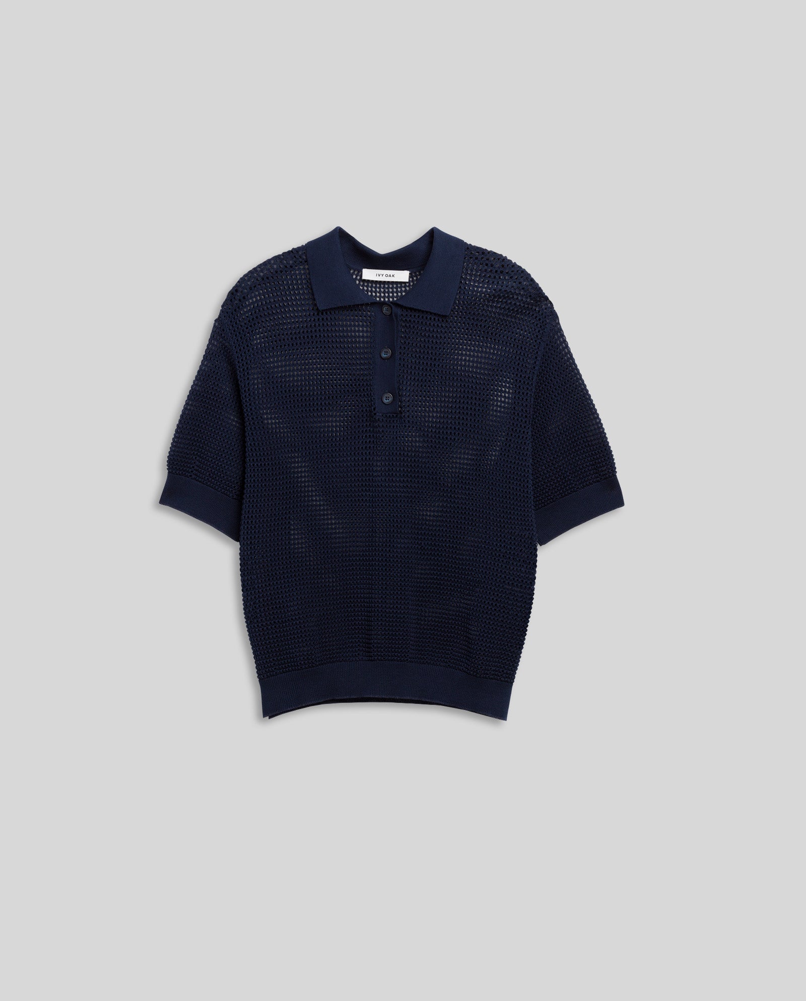 The FLO Top by IVY OAK is a navy blue short-sleeve polo shirt with three buttons and a summer open-knit, featuring a textured mesh-like pattern, shown on a gray background.