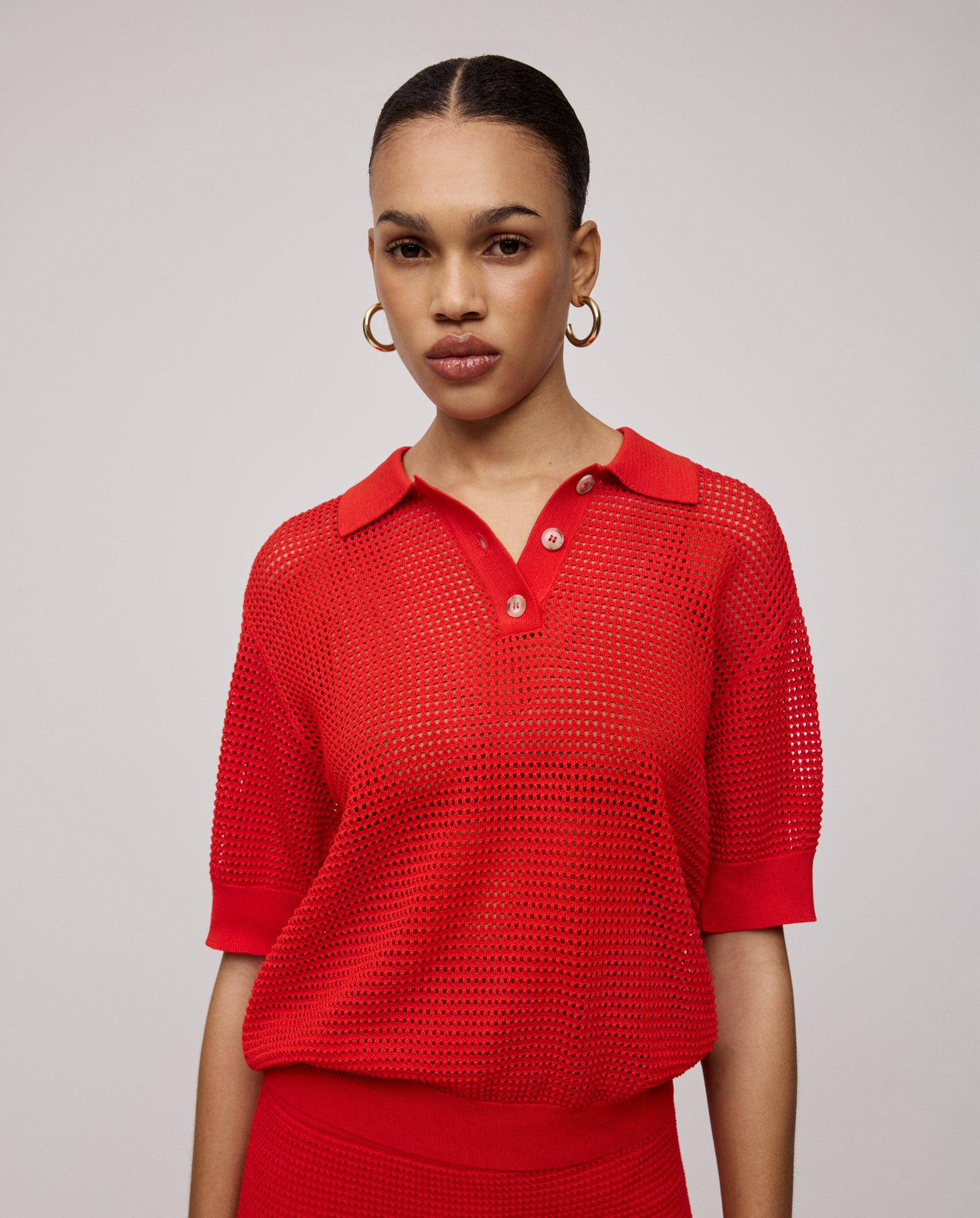 A woman wears the IVY OAK FLO Top, a vibrant red, open-knit polo shirt, styled with gold hoop earrings and standing against a plain background.