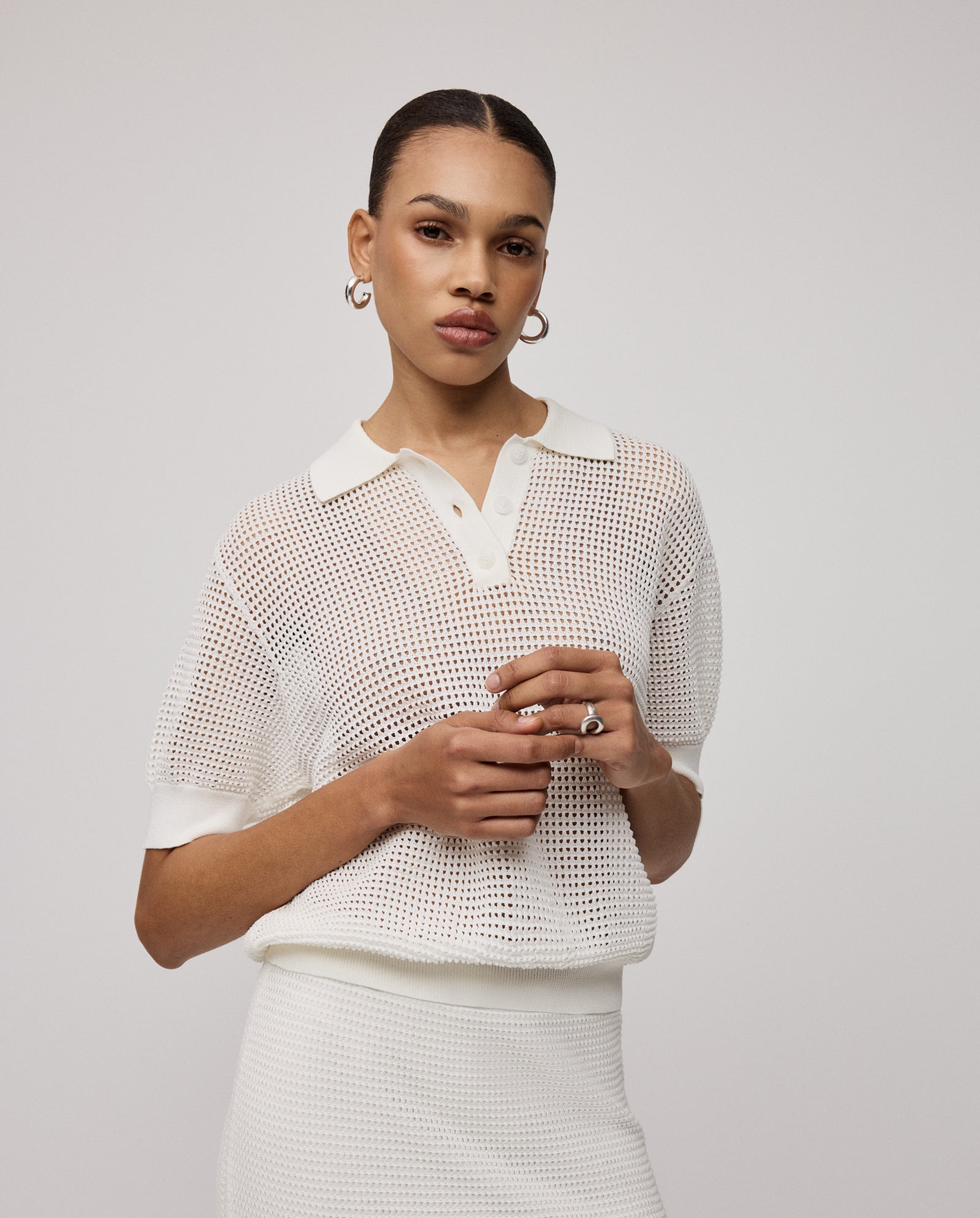 Woman wearing the IVY OAK FLO Top, a white open knit short-sleeve style, paired with a skirt and posing against a plain light background for a chic summer look.