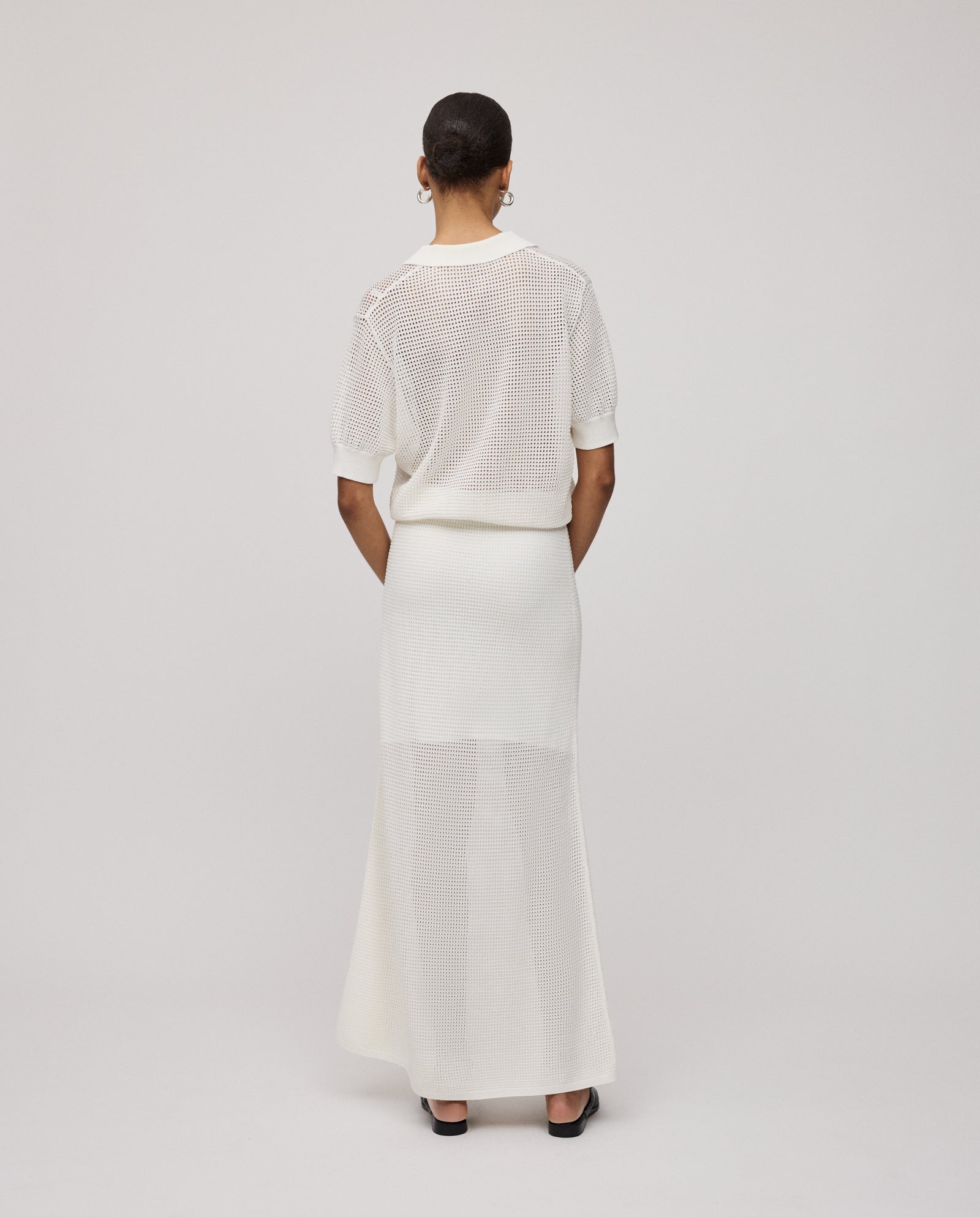 A woman stands facing away, wearing the IVY OAK FLO Top—a summerish open knit, short-sleeve polo shirt—paired with a matching long skirt and black heels.