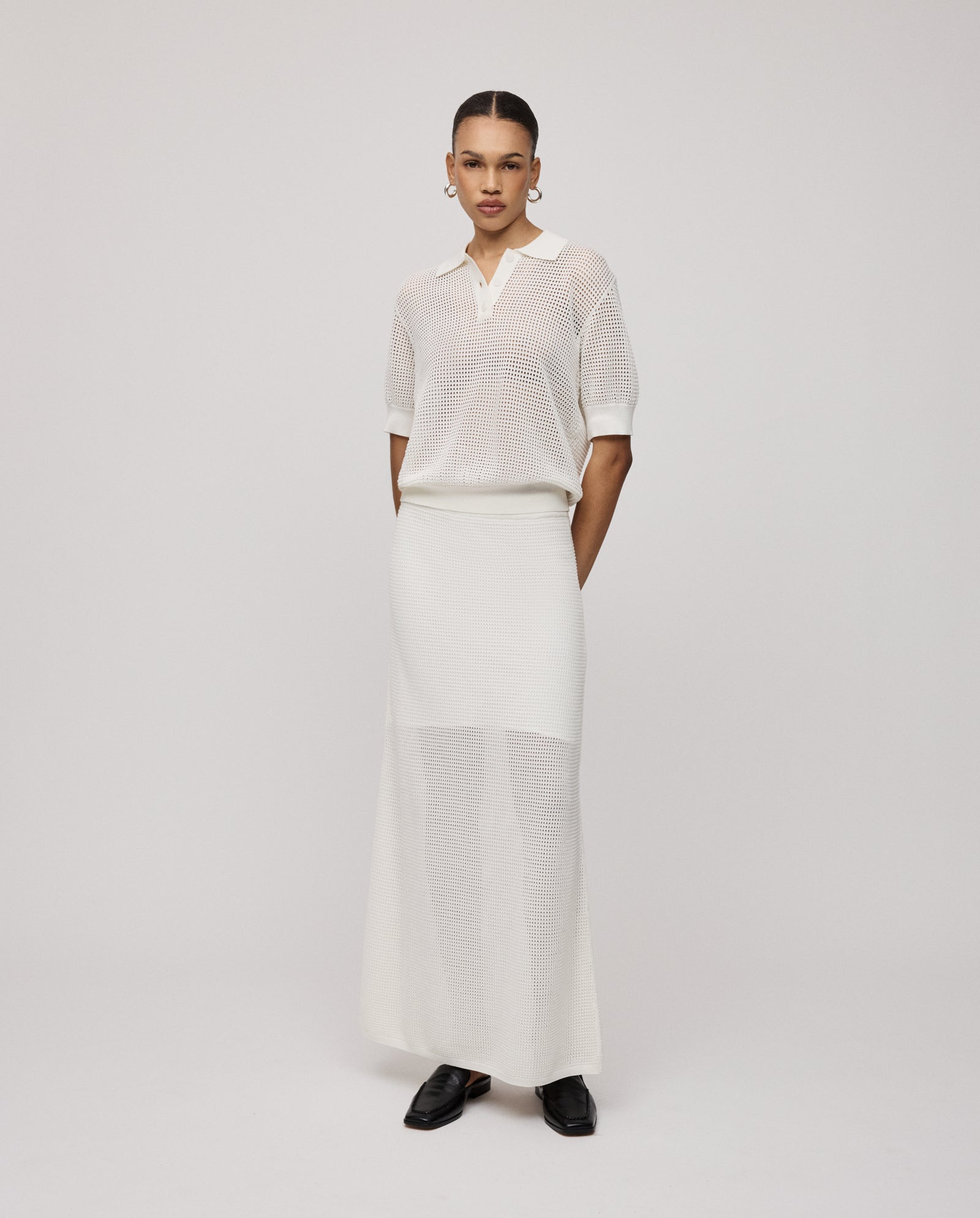 Woman wearing the IVY OAK FLO Top, a summer-inspired open knit white short-sleeve top, paired with a matching long skirt and posing against a plain light background.