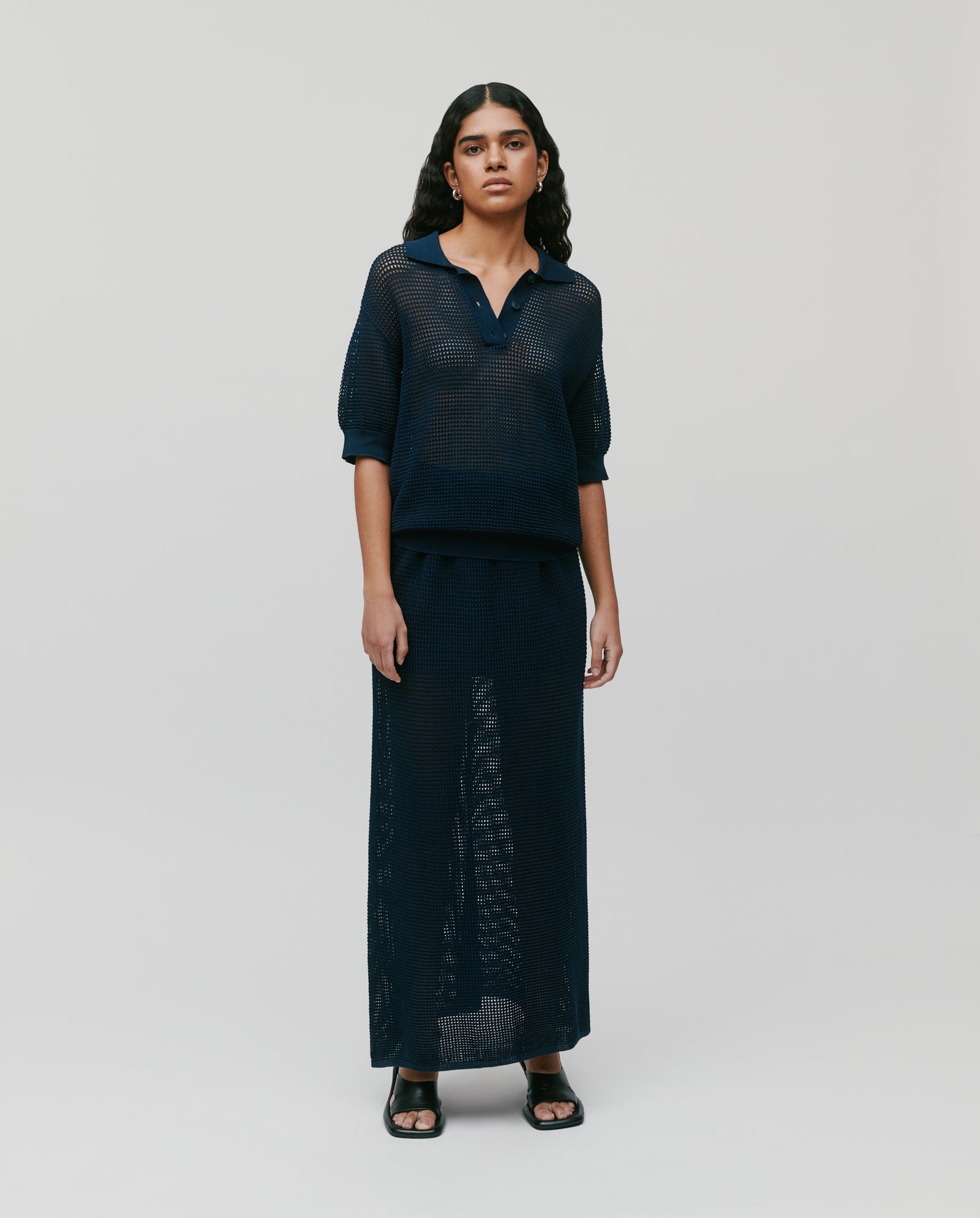 Wearing the IVY OAK FABRIZIA Skirt with a matching navy blue, sheer knit top and black sandals, this summer look is styled against a simple background.