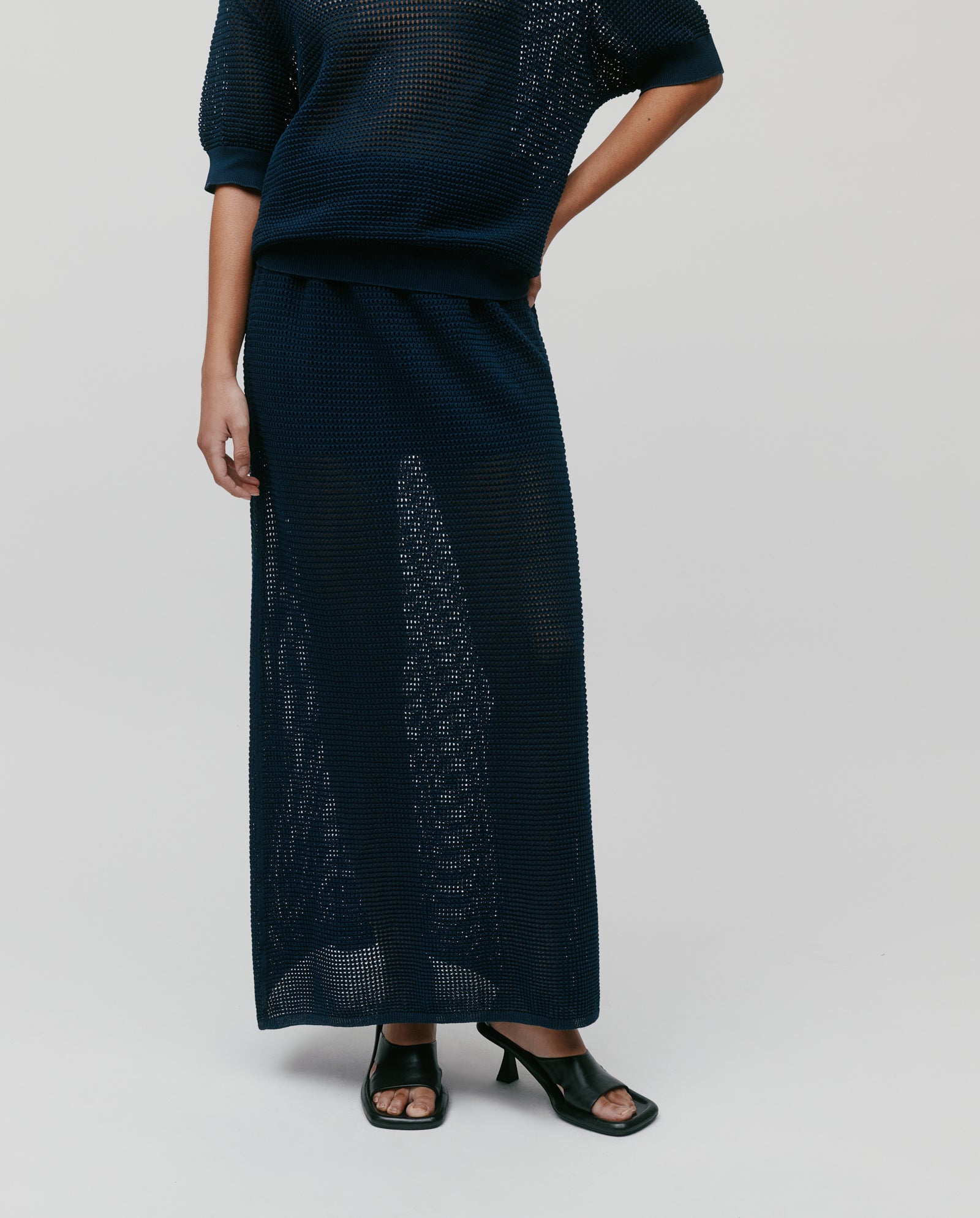 Wearing a navy blue mesh knit top and the IVY OAK FABRIZIA Skirt, paired with black heeled sandals, this look creates a summery vibe against a light background.