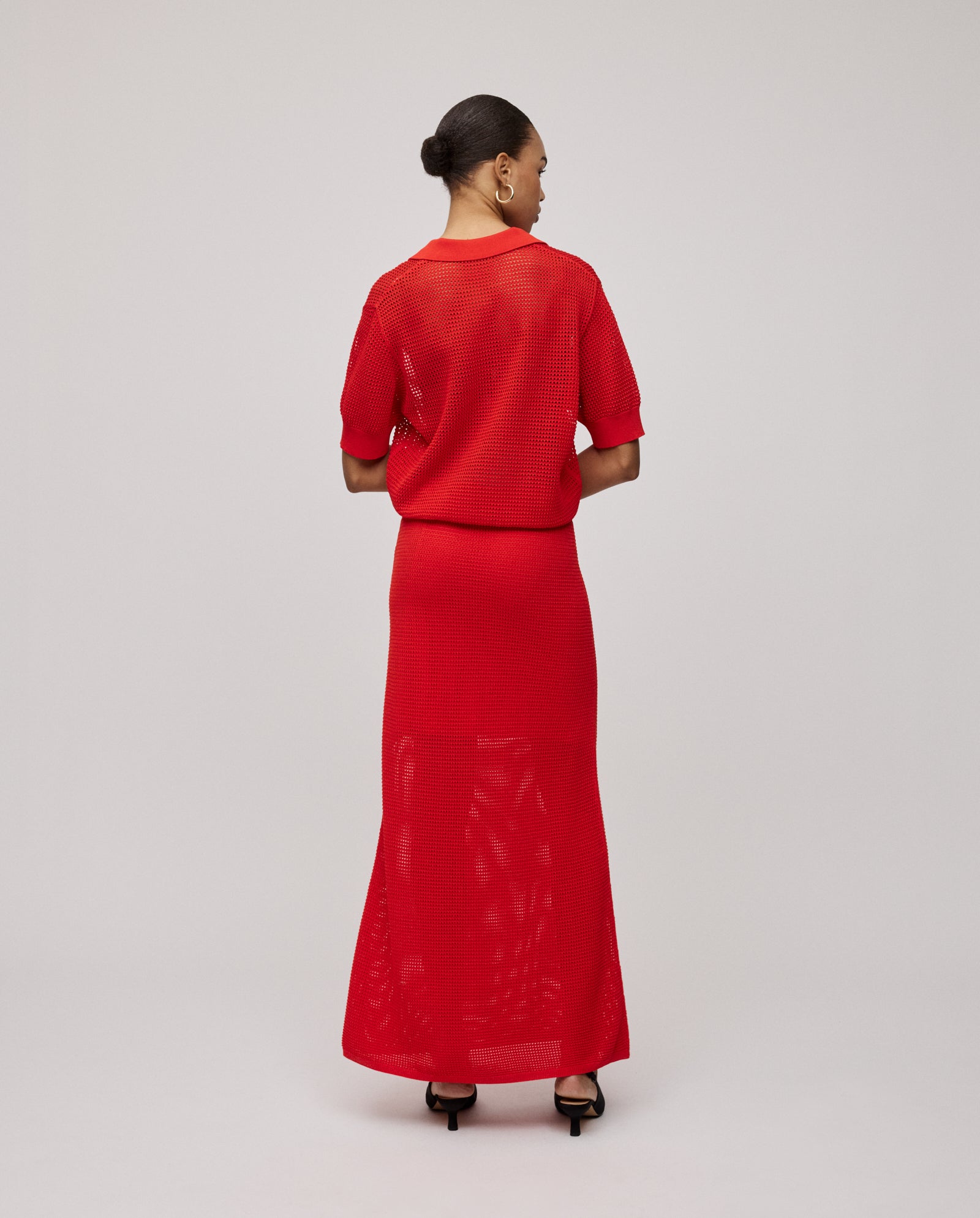 A woman faces away in an airy red IVY OAK FABRIZIA Skirt and matching top, her hair in a bun and black heels finishing the look. The light A-line and open knit give it effortless summer elegance.