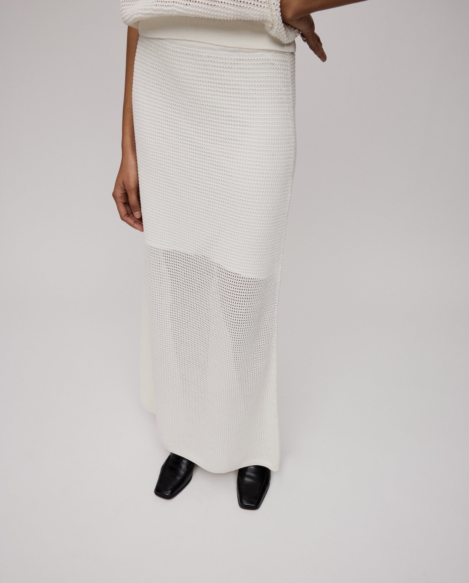 A person wears the IVY OAK FABRIZIA Skirt, a long white open-knit piece with an A-line silhouette, styled with black square-toe shoes and one arm bent at the elbow.