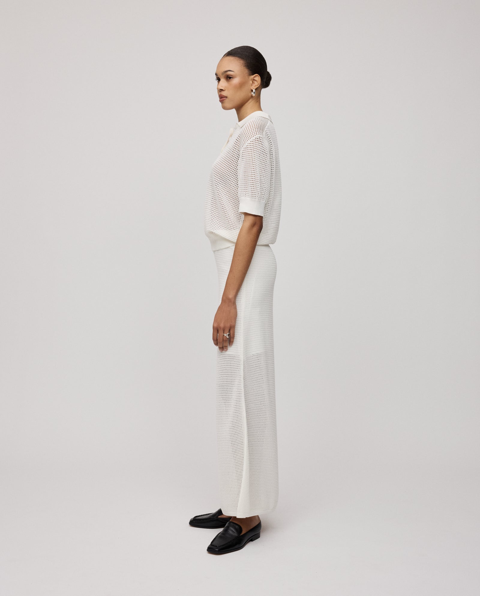 A woman stands sideways in black shoes, wearing a white blouse and the IVY OAK FABRIZIA Skirt—a summer knit with an A-line silhouette—set against a plain background.