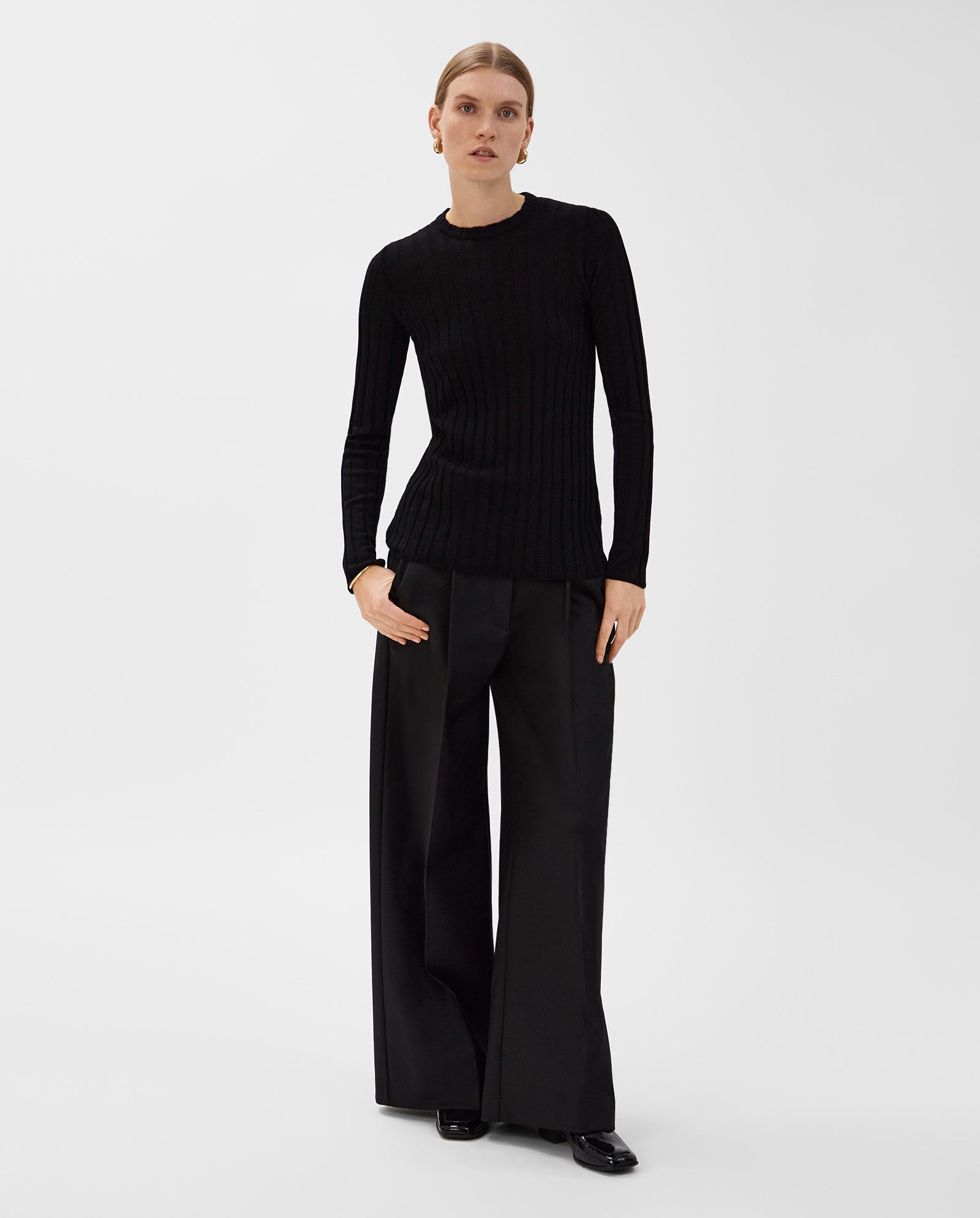 A woman poses in the IVY OAK KACY ANN Knittop, a precise-fit black Merino rib sweater, paired with wide-leg black trousers against a plain white background.